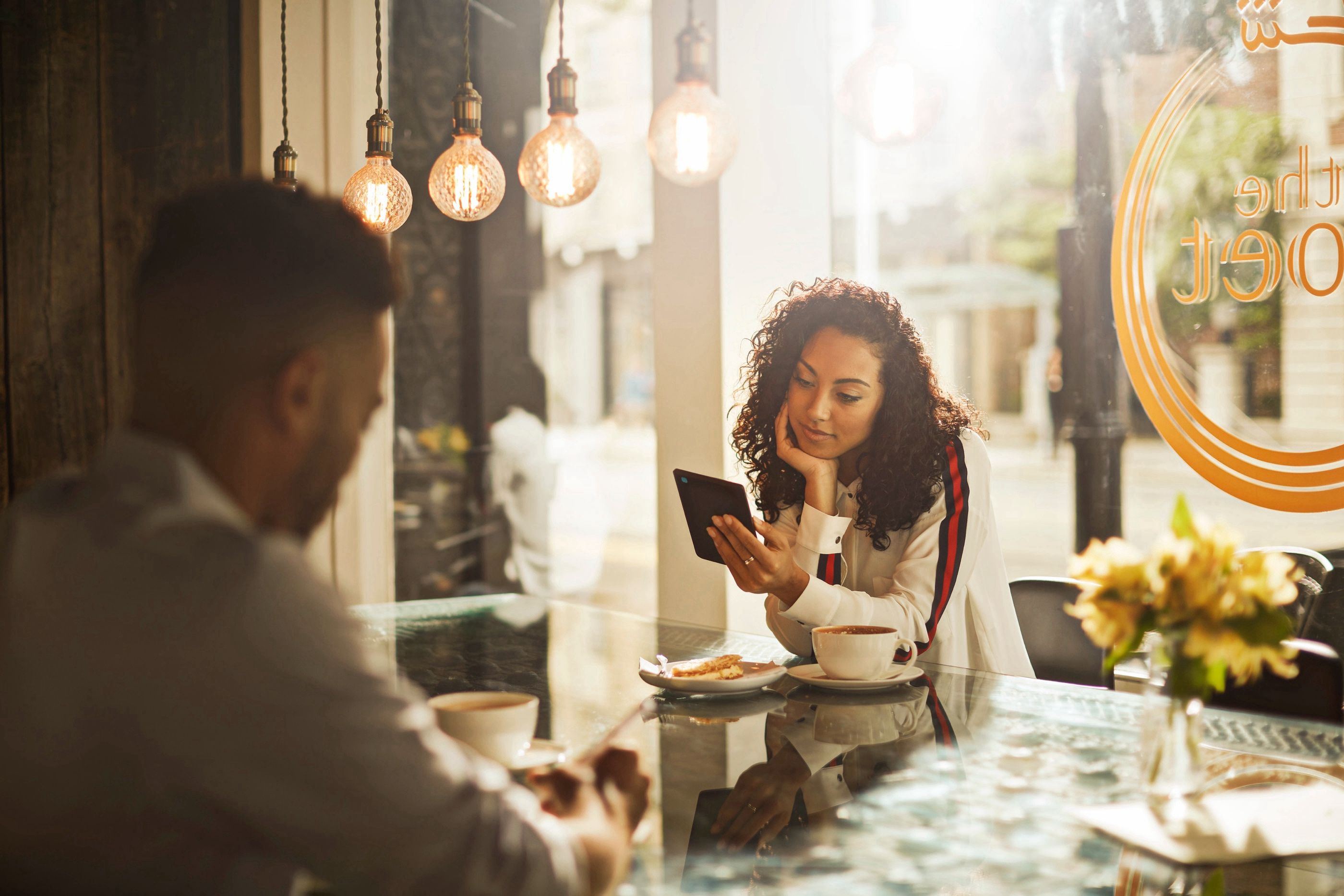 Woman with curly hair looks at a tablet at a cafe table across from a man, coffee cups and pastries nearby.