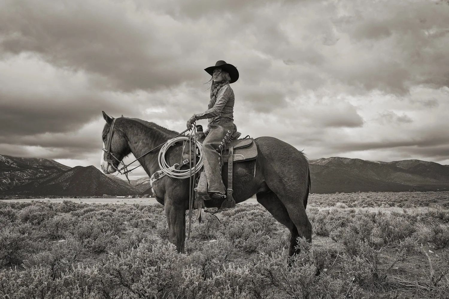Black-and-white photo of a woman in a cowboy hat riding a horse through sagebrush, mountains in the background.