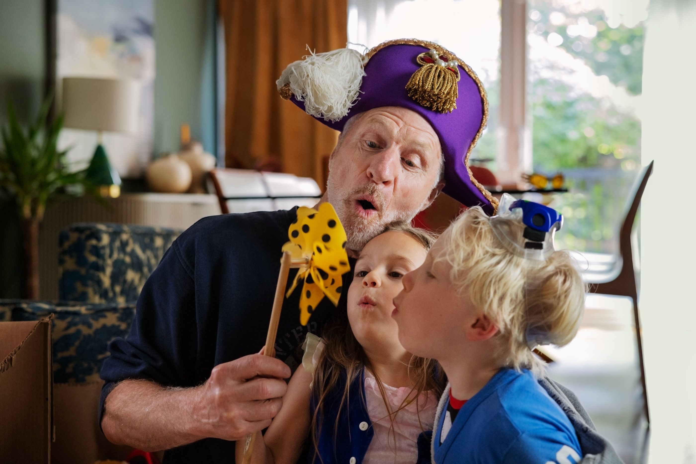Man wearing a purple pirate hat plays with two young children in a living room.