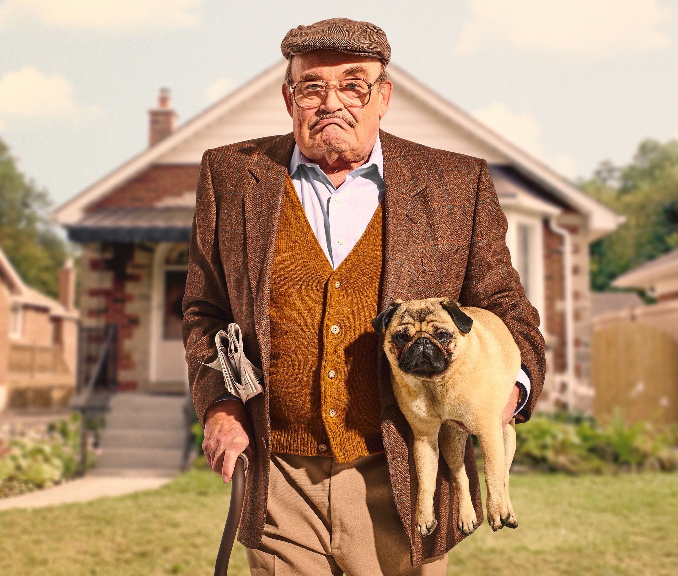 Older man in a tweed jacket and cap carries a pug under his arm while walking with a cane in a yard.