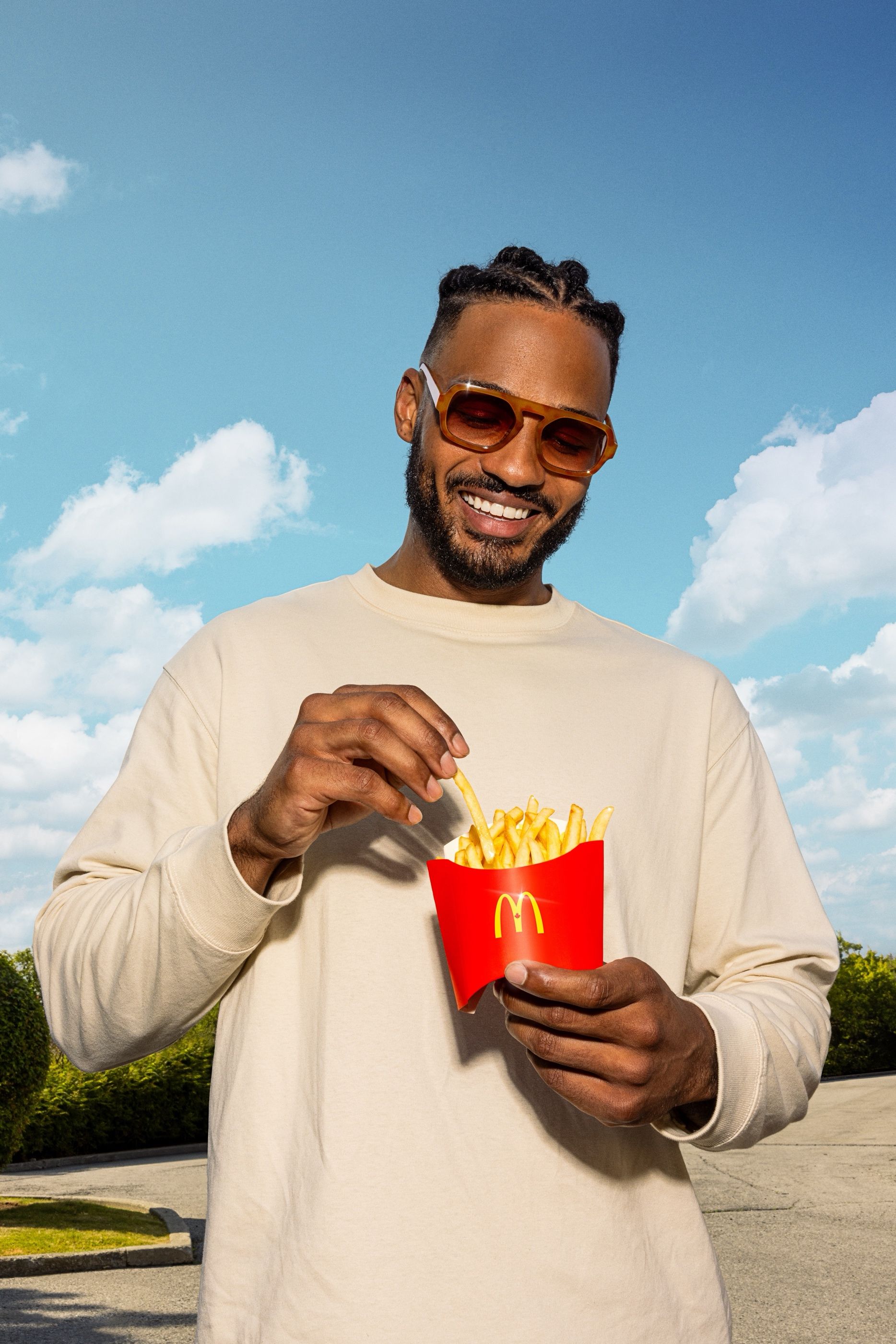 Man in sunglasses eating fries from a McDonald's fry box outdoors under a blue sky.