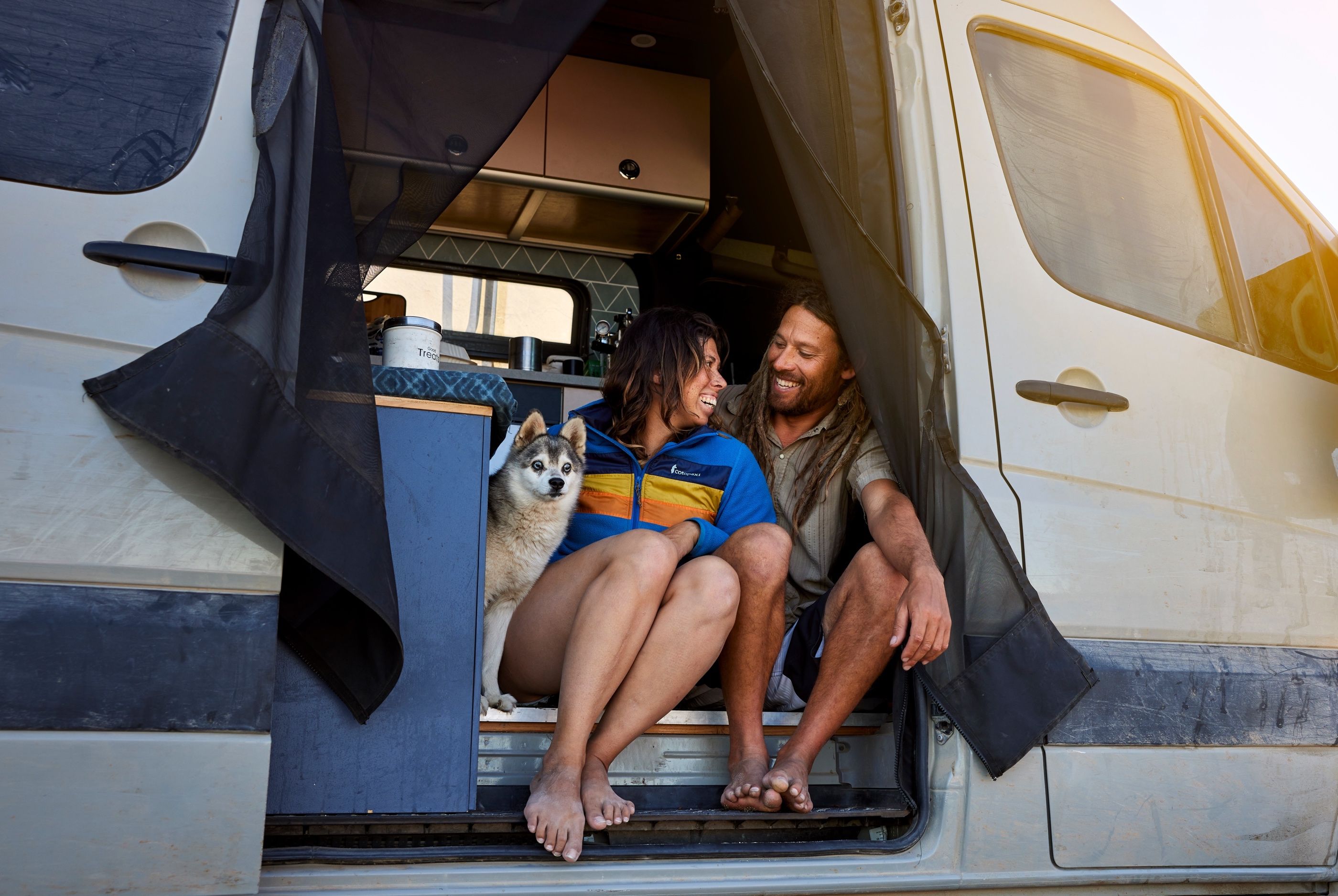 Couple sitting in the open side door of a camper van, laughing together with a husky dog beside them.