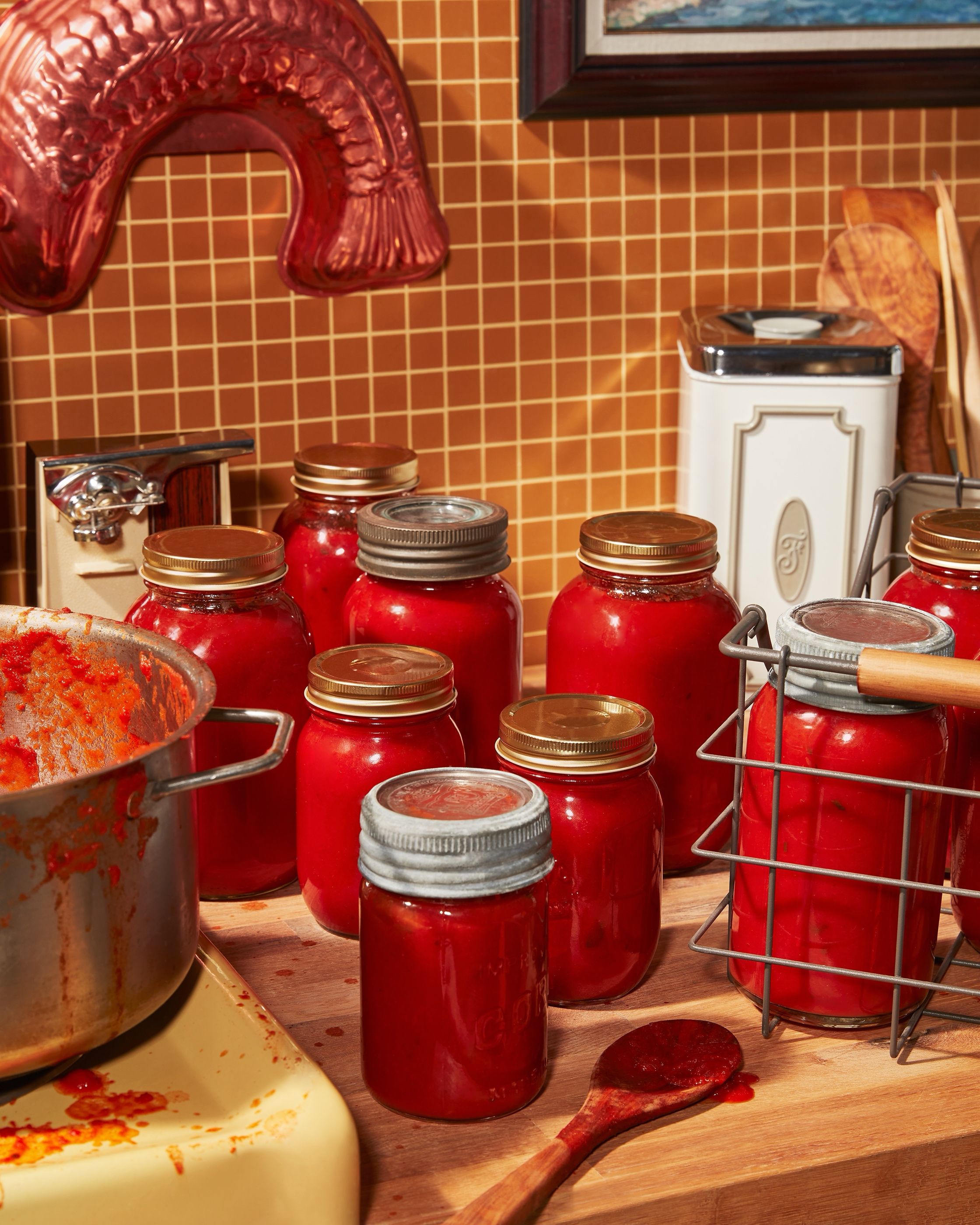 Jars filled with red tomato sauce lined up on a wood counter next to a large pot, part of a home-canning setup.