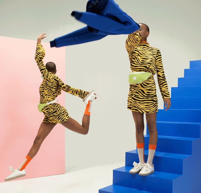 Two women in tiger-print outfits on a blue staircase set, one leaping, one holding a blue jacket.