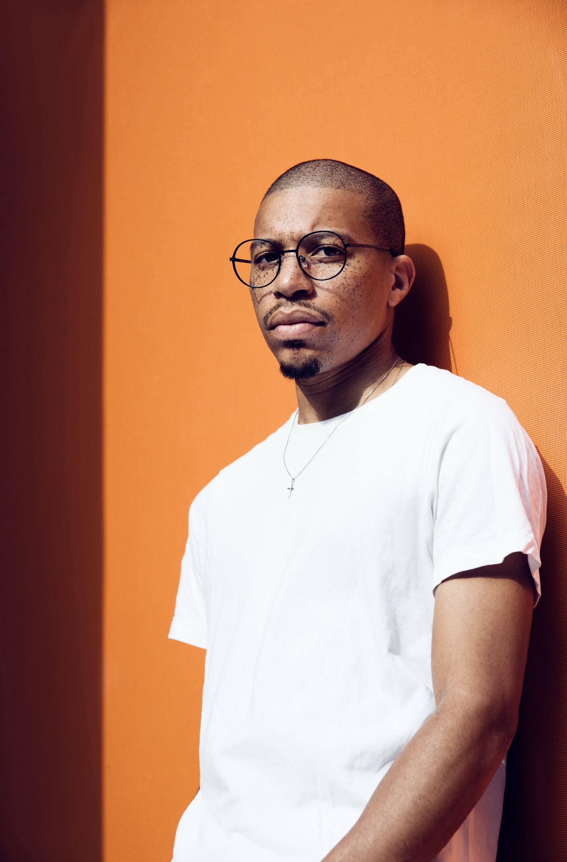 Man with round glasses and a short beard leans against an orange wall, wearing a white t-shirt and a silver cross necklace.