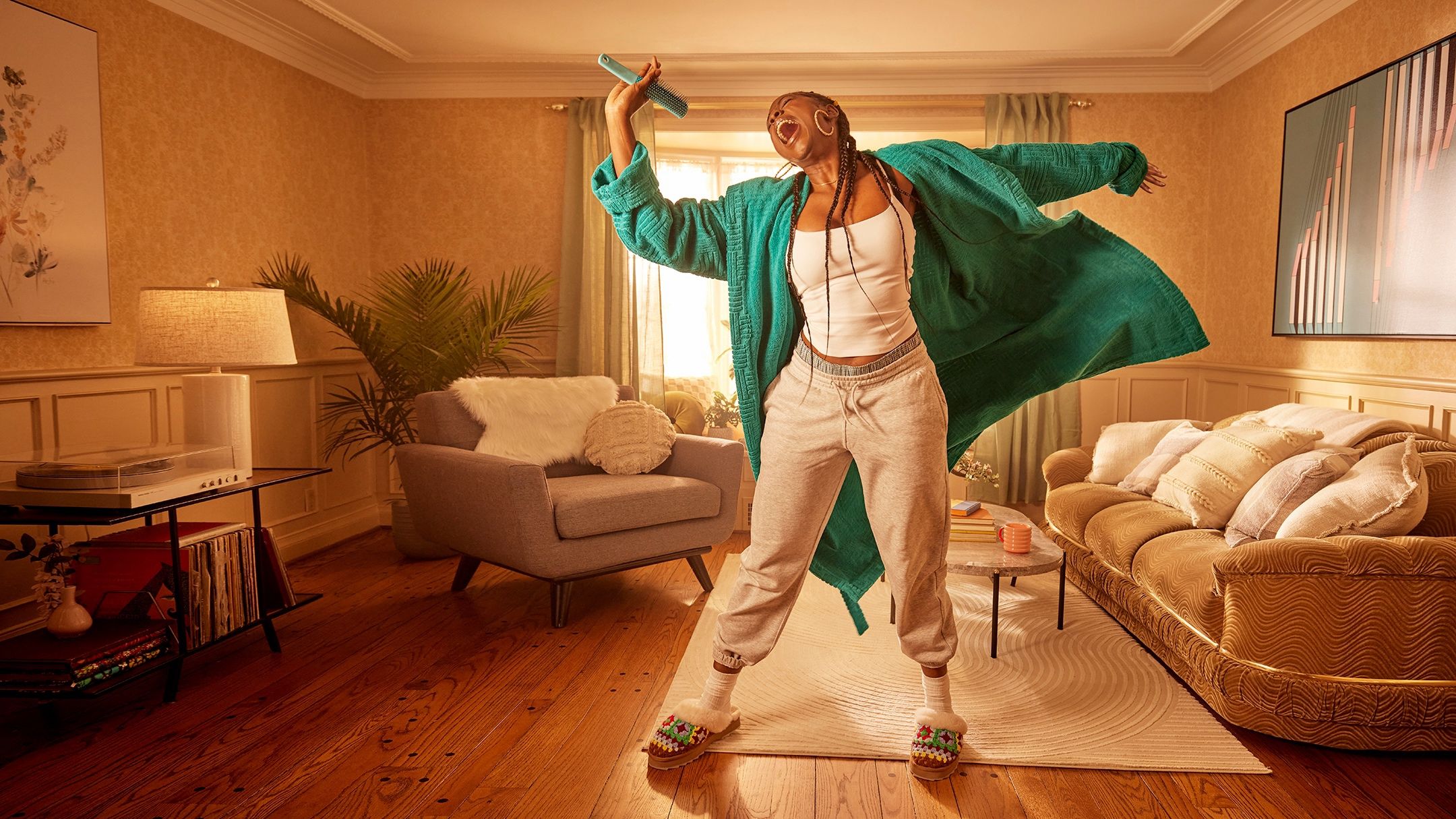 Woman in a green robe jumps and sings into a hairbrush in a warmly lit living room with a record player.