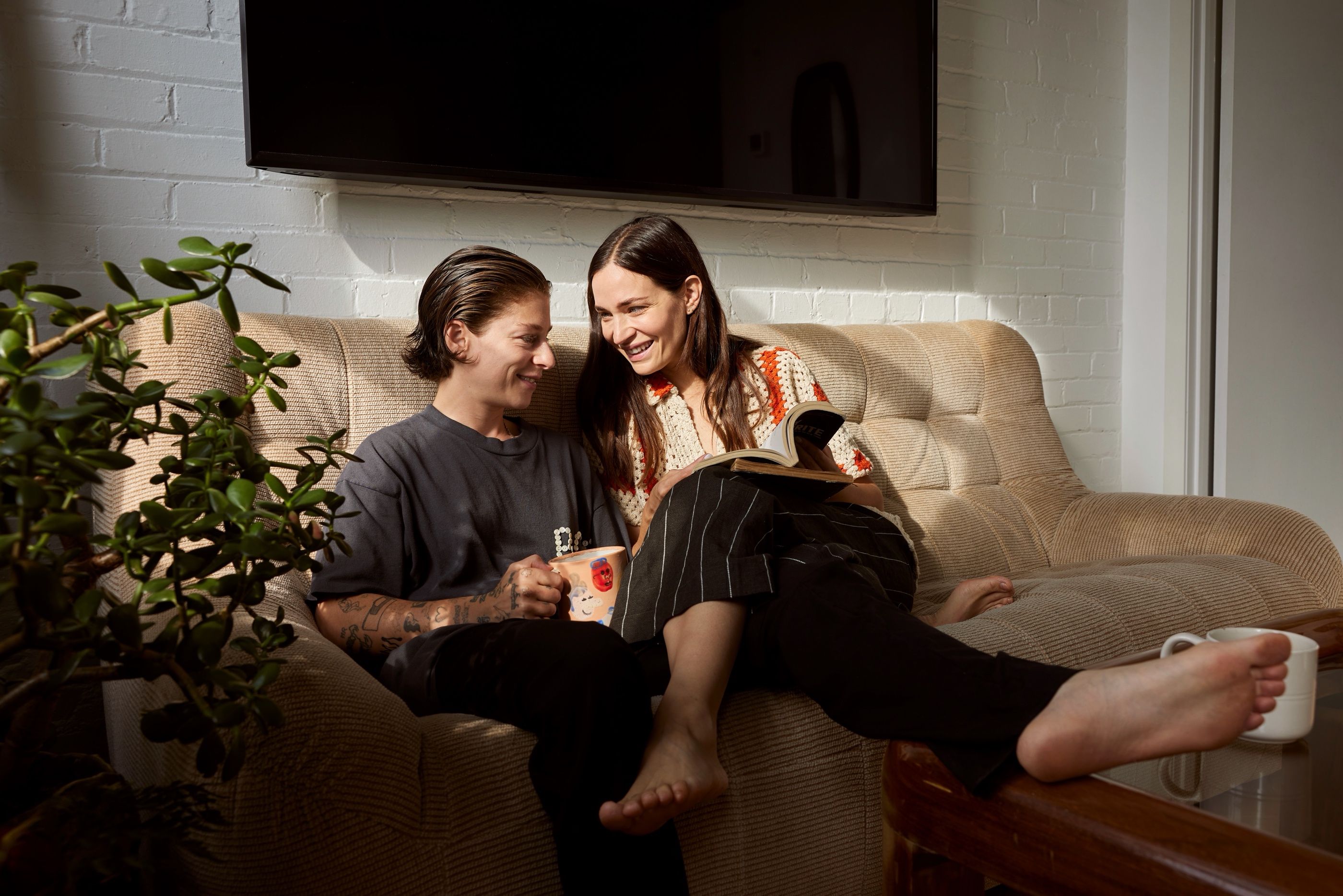 Couple sitting together on a couch at home, one holding a mug and a book, feet resting on a coffee table.