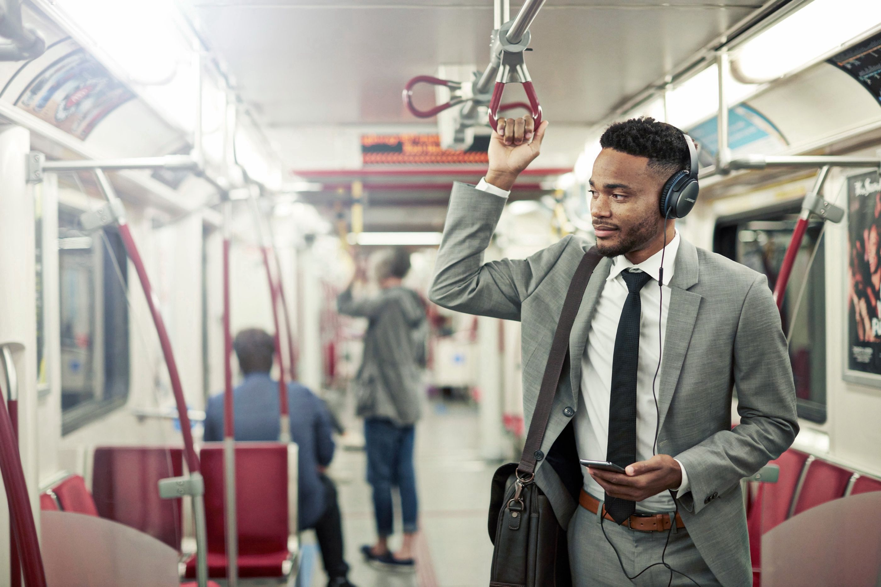Man in a suit stands on a subway train holding an overhead handle and a phone, wearing headphones.