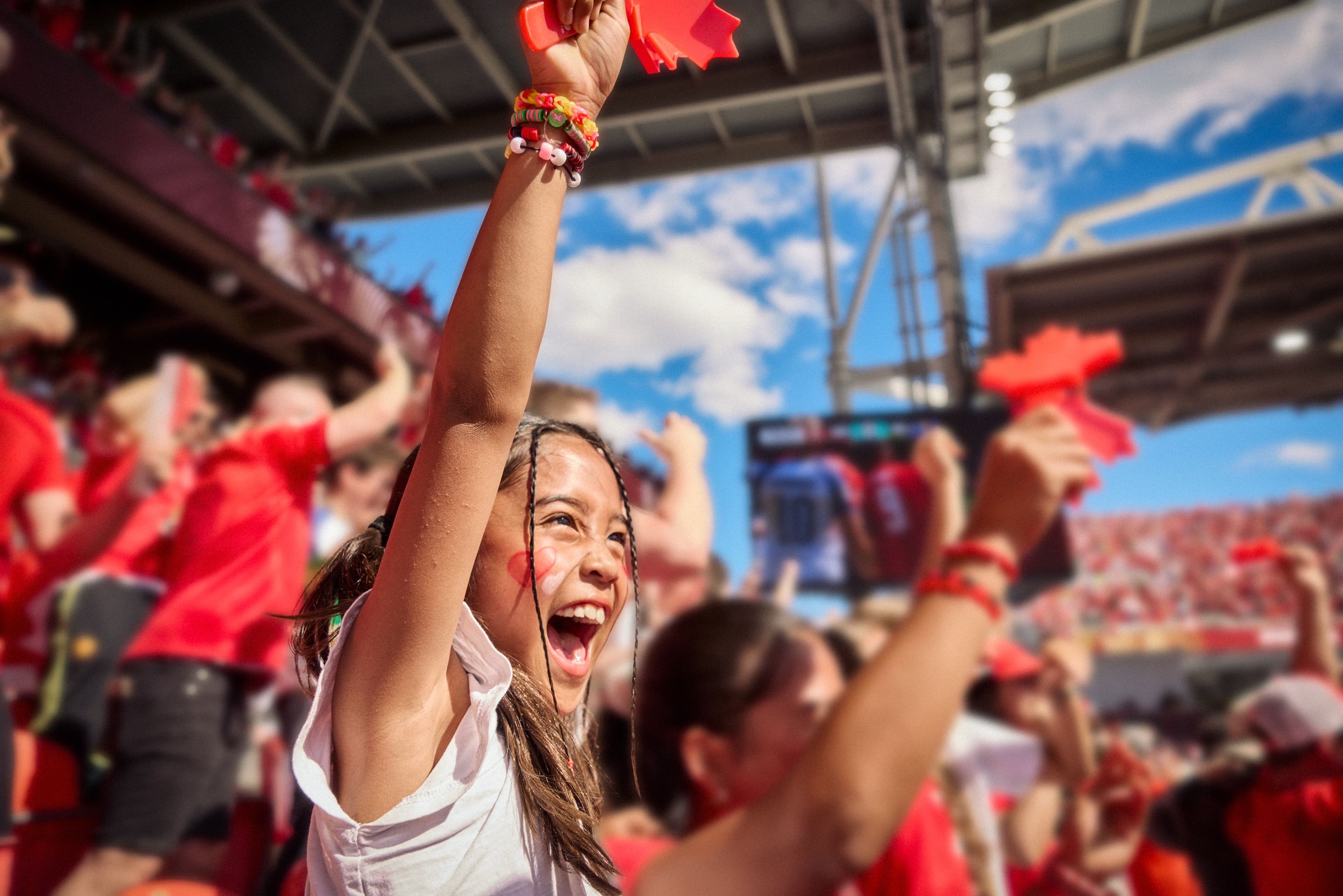 Girl cheers with a raised fist in a crowded outdoor stadium, wearing red face paint.