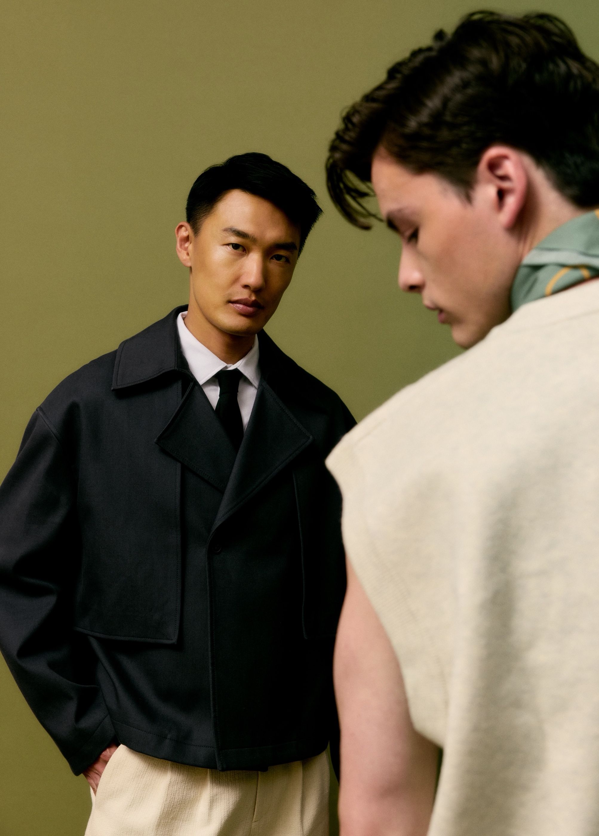 Two men in tailored jackets and knitwear standing close together against an olive green background.