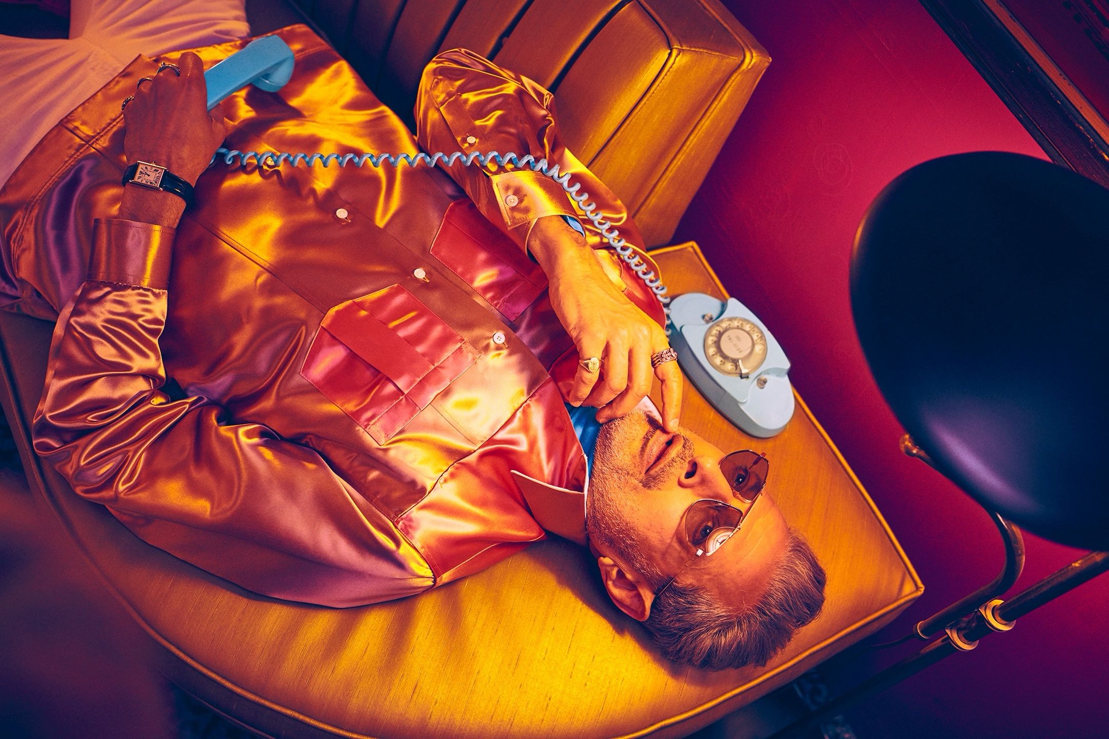 Man in a satin jacket and sunglasses reclines on a table, talking on a vintage rotary phone in a red-lit room.