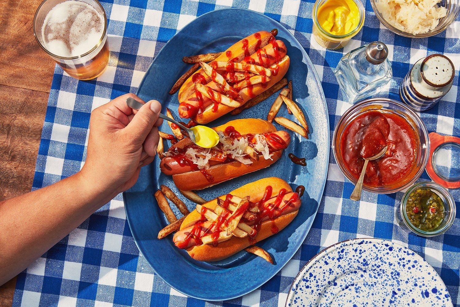 A hand adds mustard to hot dogs topped with ketchup and fries on a blue plate, with condiments alongside.