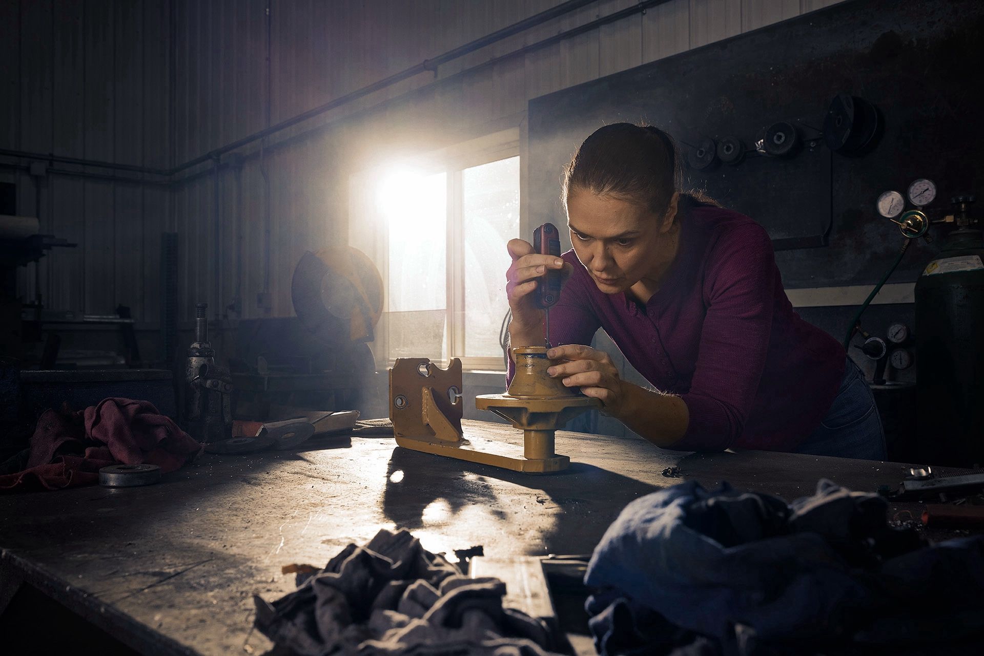 Woman leans over a workbench using a hand tool on a piece of yellow machinery in a dim workshop.