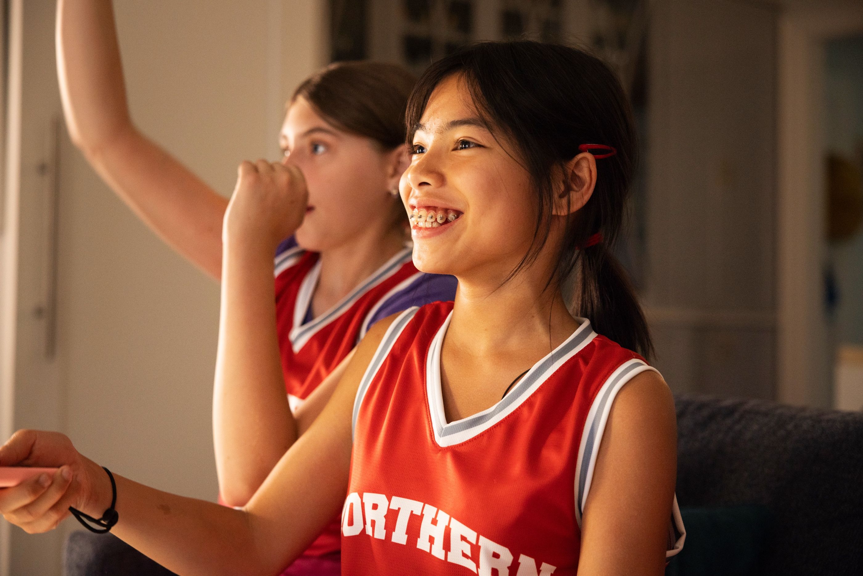 Two girls in red "Northern" basketball jerseys smile indoors, one wearing braces.