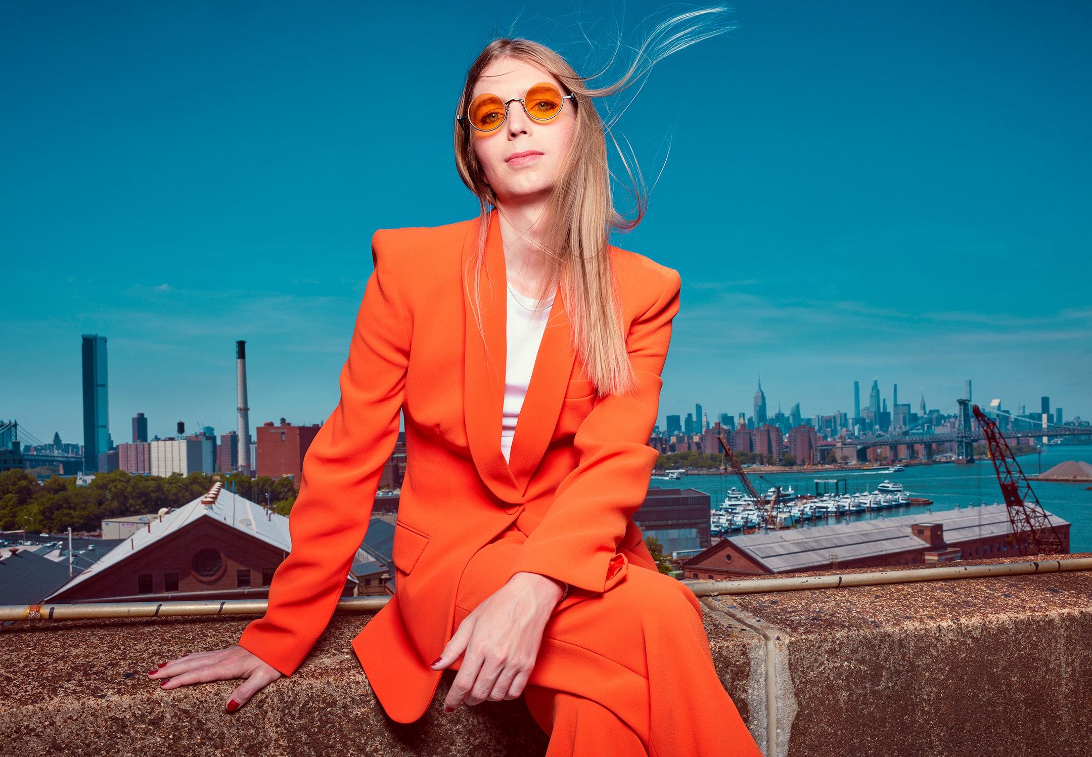 Woman in an orange suit and orange-tinted sunglasses sits on a rooftop ledge with a city skyline behind her.