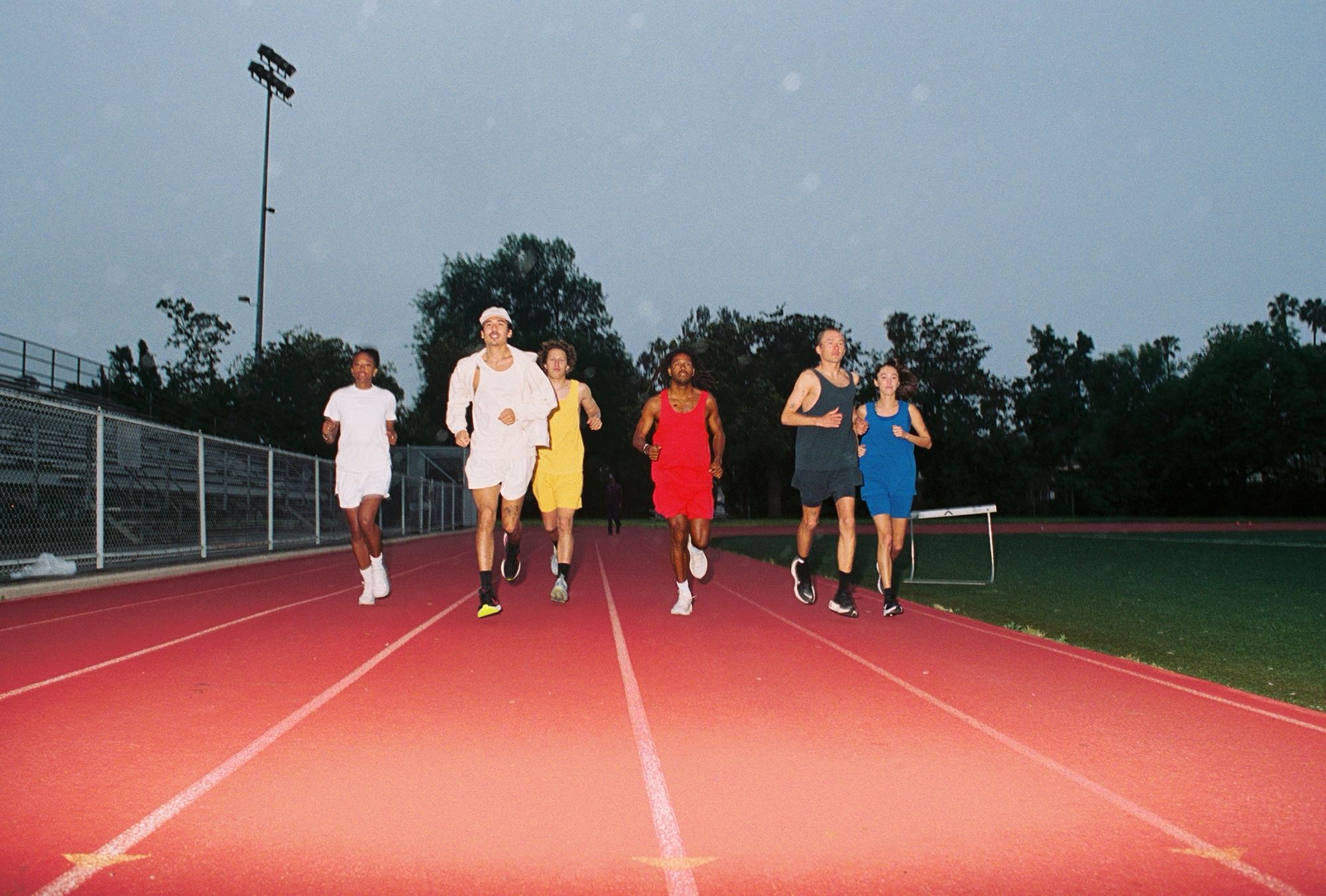 Group of six people jogging together on an outdoor running track at dusk.