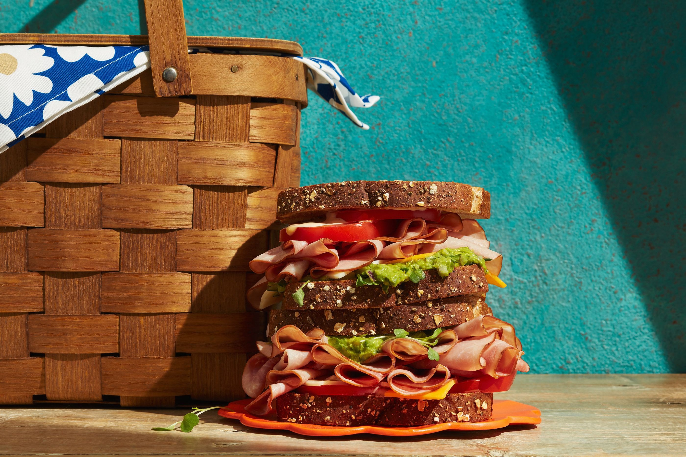 A tall deli sandwich with layers of ham, cheese, tomato and avocado spread on dark bread, beside a wicker picnic basket.