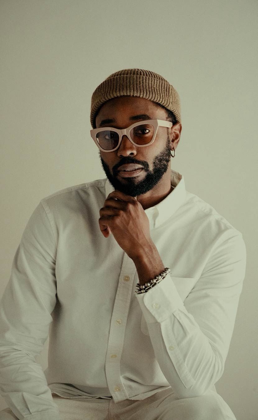 Man wearing a beige beanie and pink-tinted sunglasses, hand at chin, white button-down shirt.