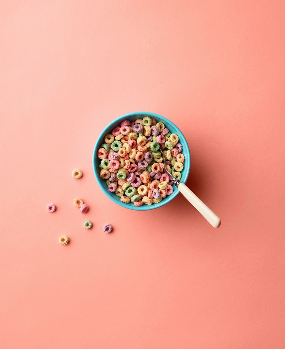 A blue bowl of colorful fruit loop cereal with a spoon, scattered loops on a pink background.