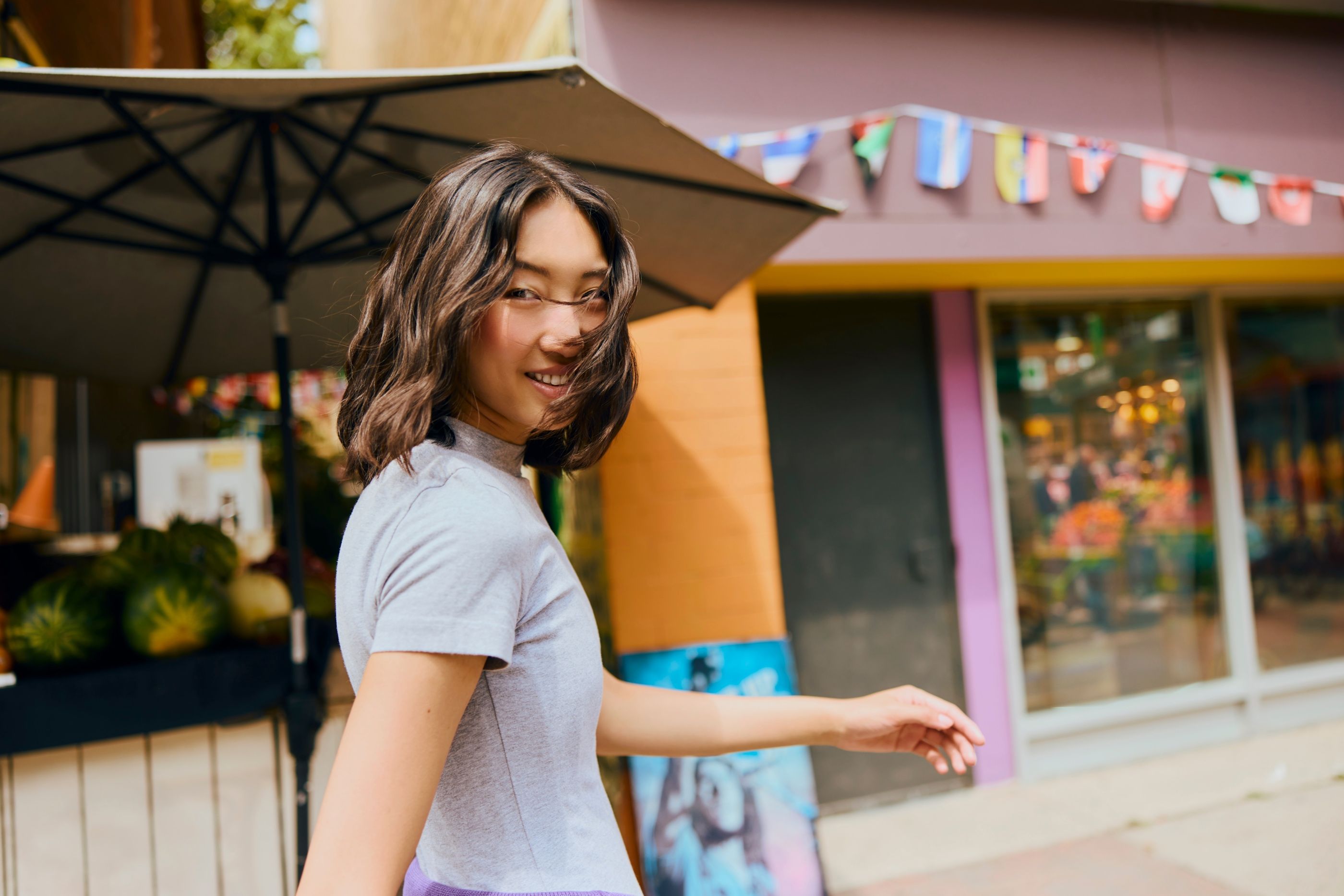 Woman with wavy chin-length hair looks back over her shoulder while walking past a market stall with flags overhead.