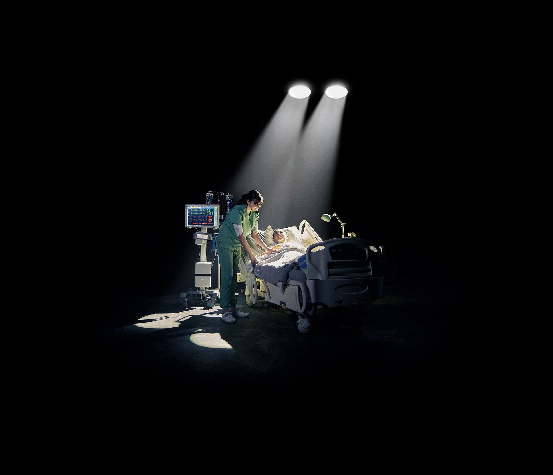 Health care worker in green scrubs attends to an older patient in a hospital bed, dramatically lit against black.