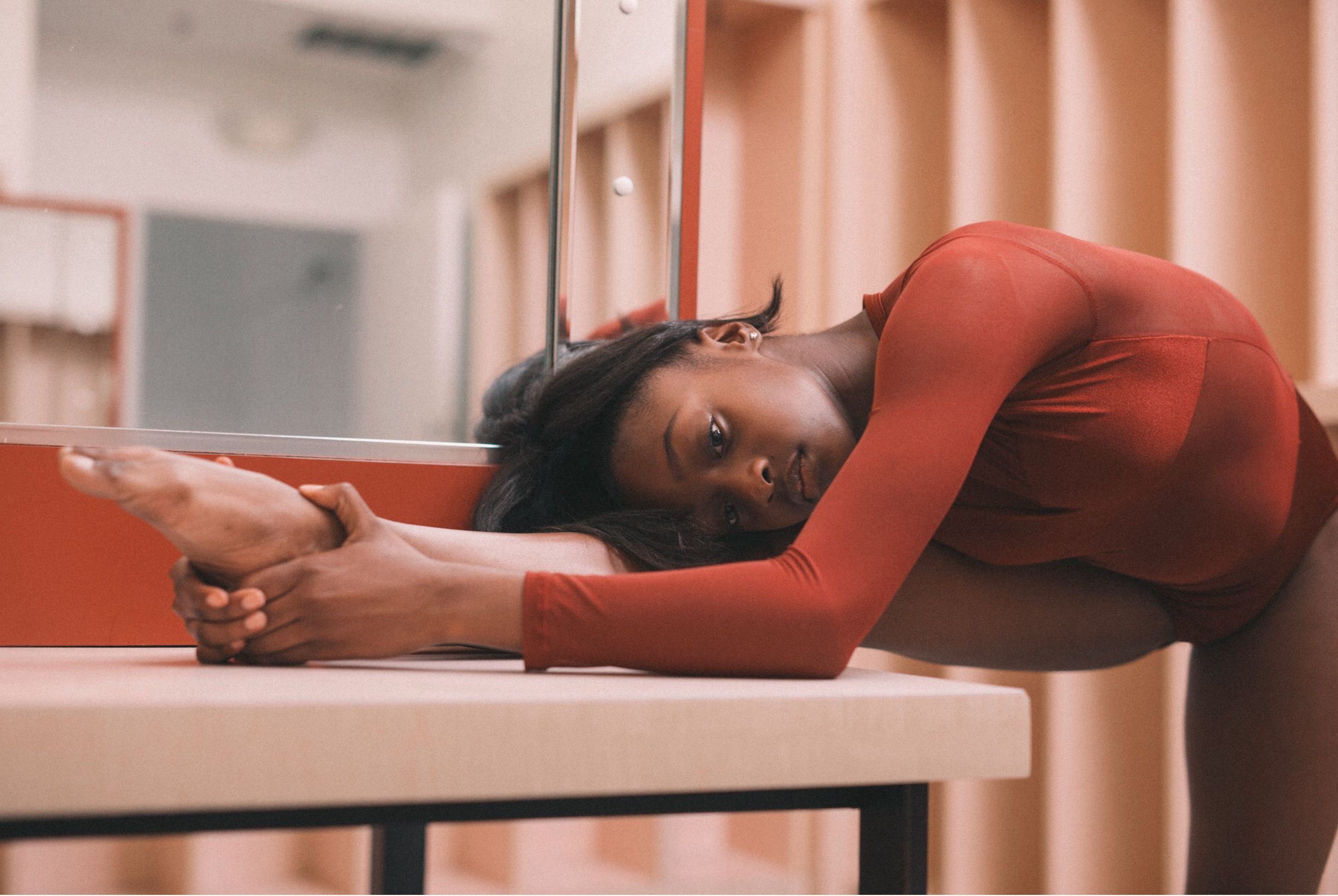 Woman in a red long-sleeve leotard stretches over a bench in a mirrored studio.