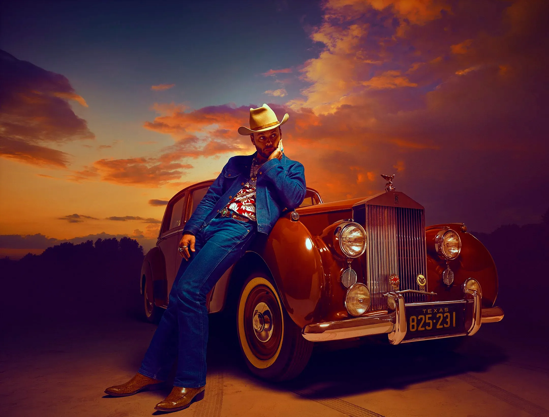 Man in a cowboy hat and denim jacket leans against a vintage convertible car at sunset.