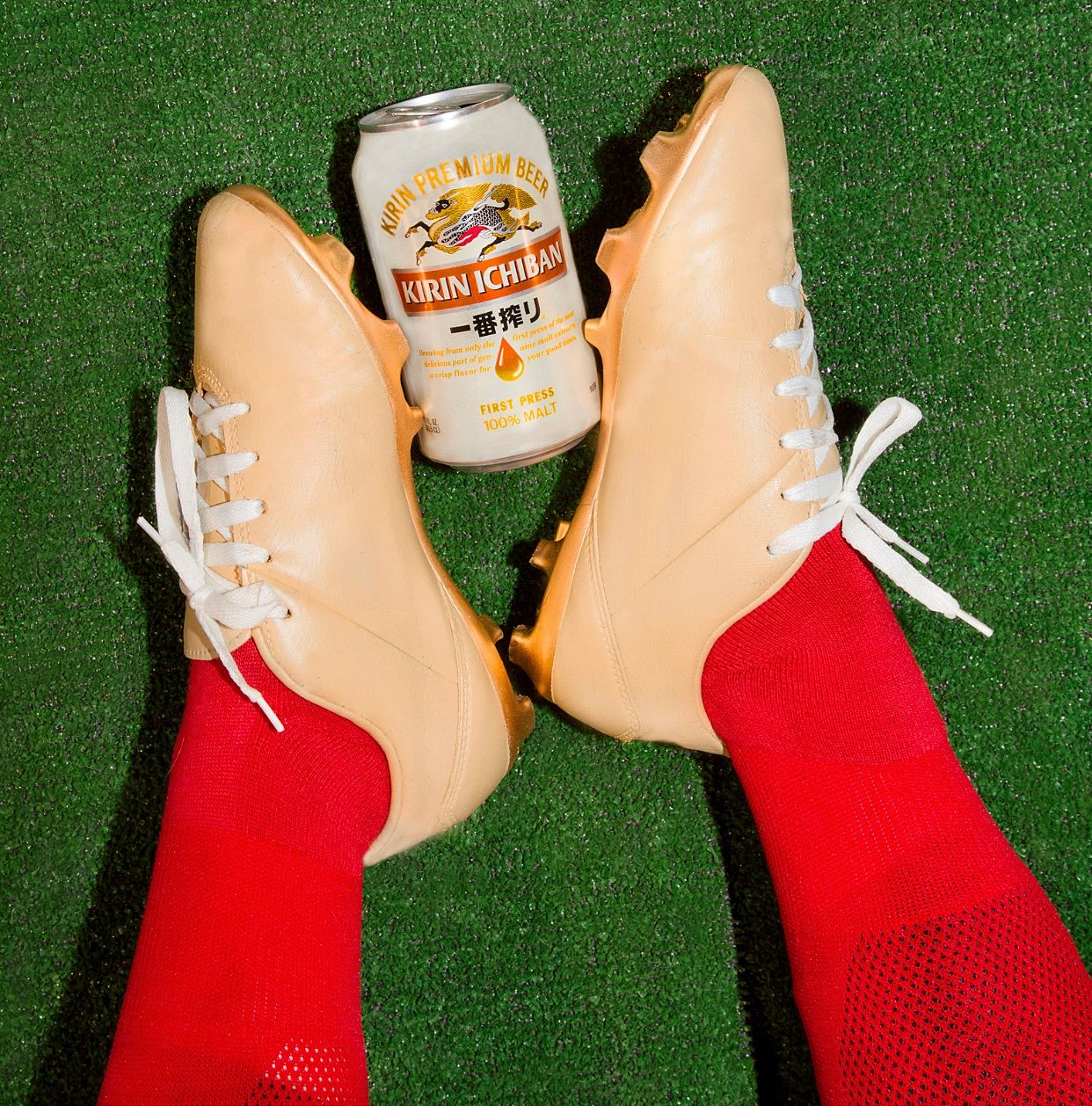Gold cleats with red socks next to a can of Kirin Ichiban beer on artificial turf.