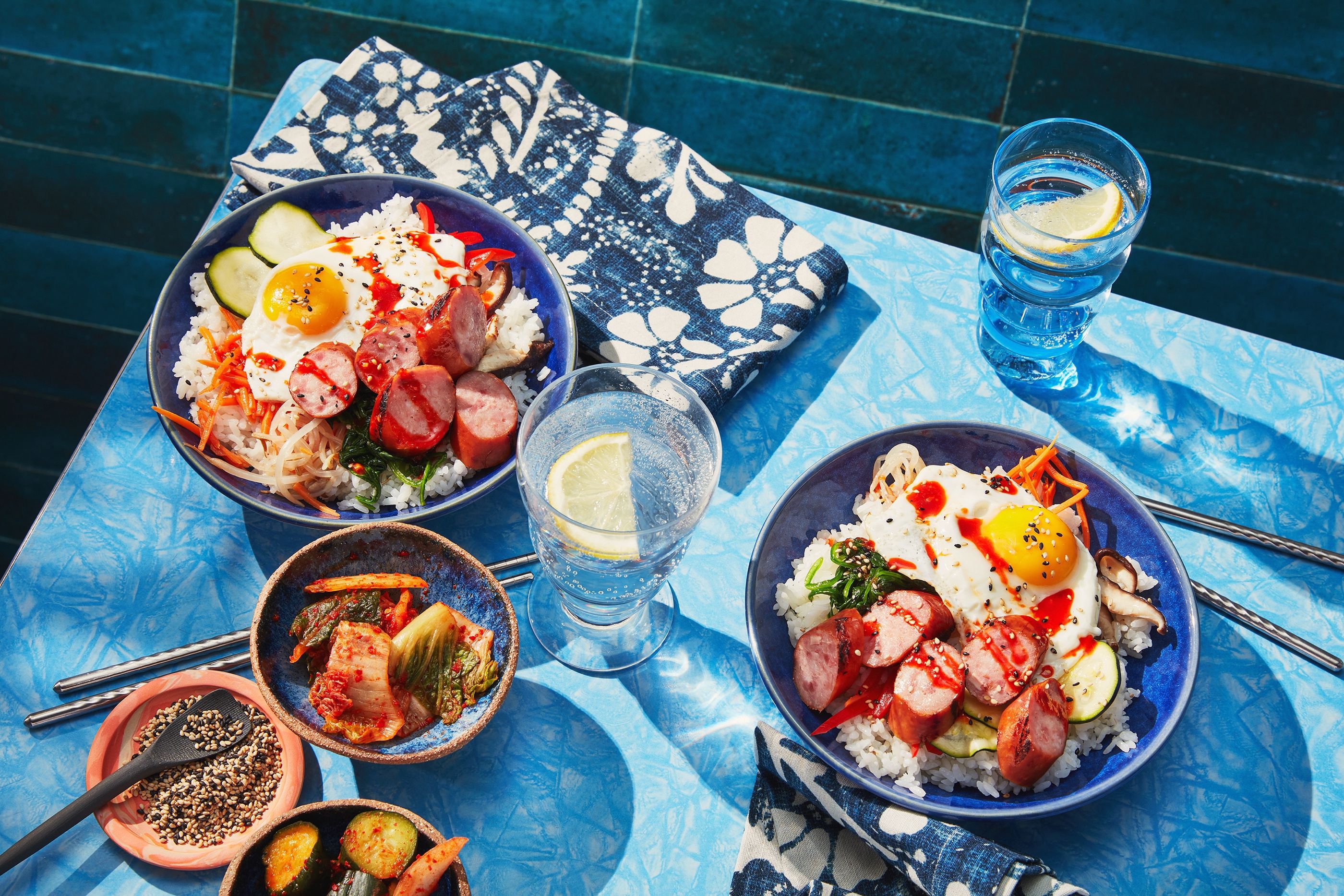 Two rice bowls topped with sliced sausage, a fried egg and vegetables, served with kimchi and chopsticks on a blue table.