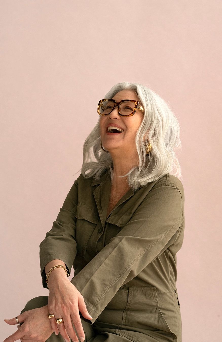 Older woman with gray hair laughing, wearing tortoiseshell glasses and an olive green jumpsuit.