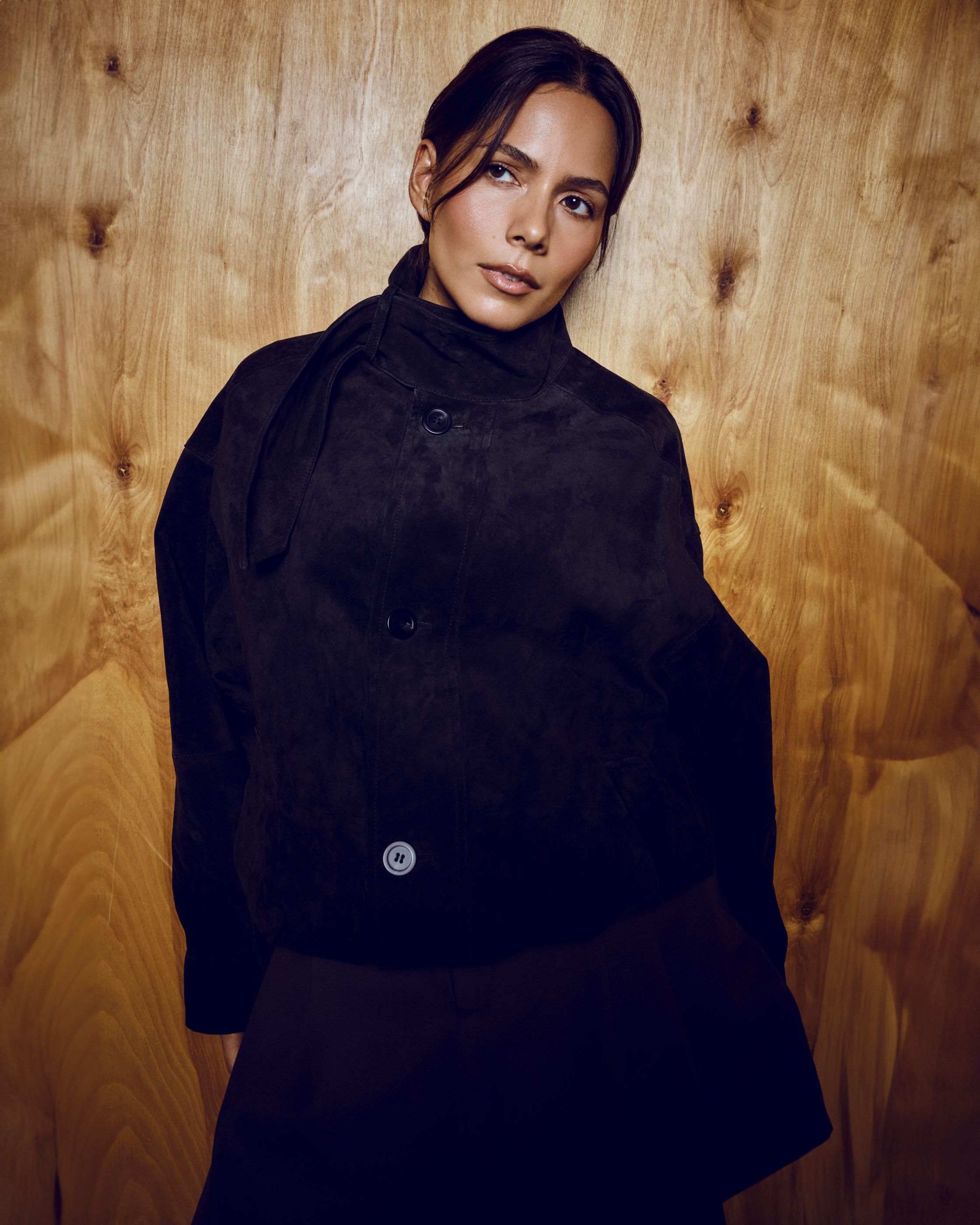Woman in a black suede jacket with a high tie collar stands against a warm wood-paneled wall.