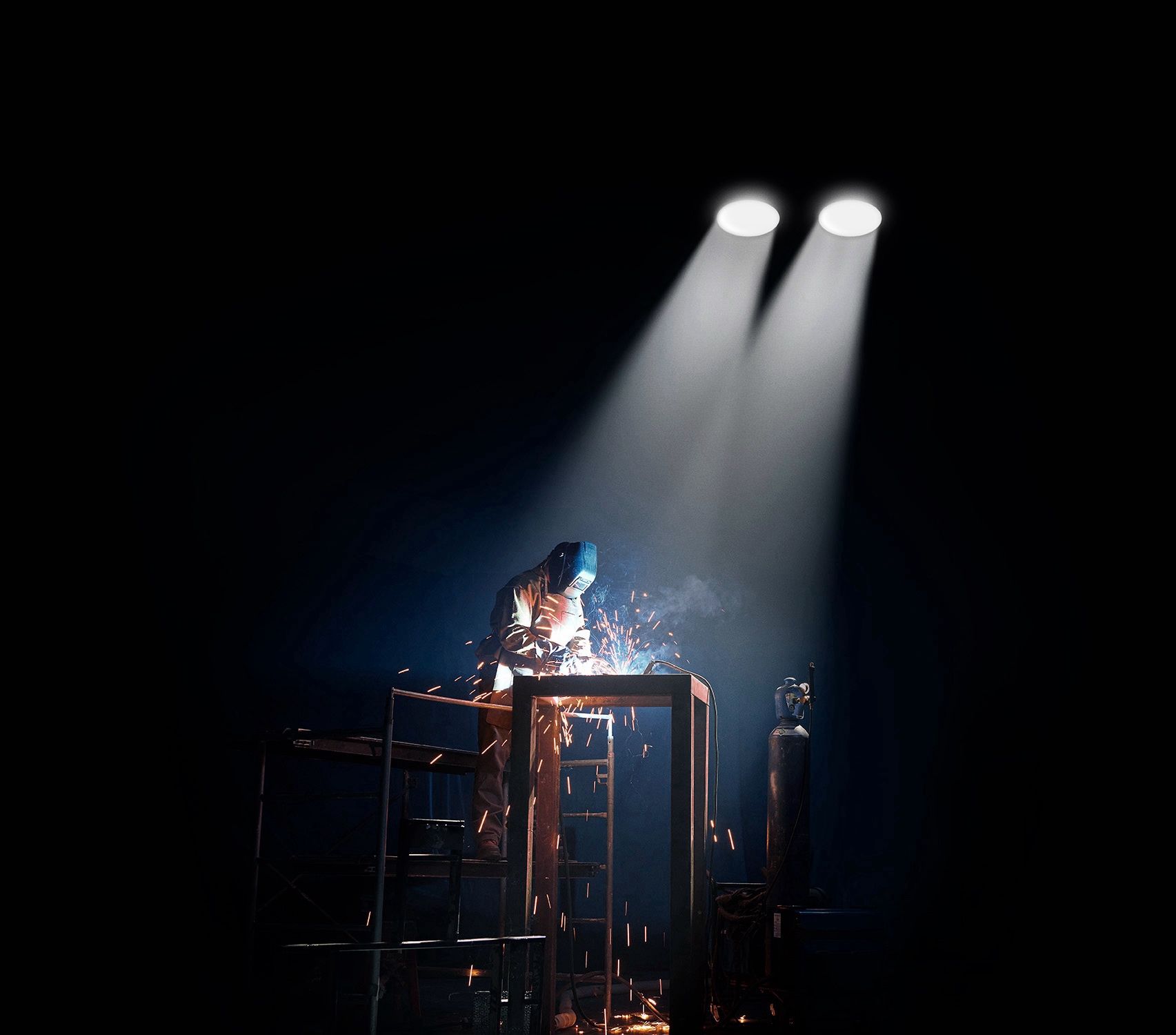 Welder in protective gear works on a metal frame, sparks flying, dramatically lit against a dark background.