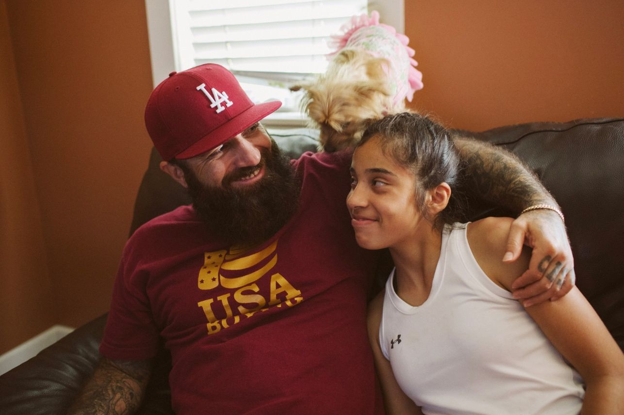 Man in a red LA cap and "USA Boxing" shirt hugs a girl on a couch, with a small dog nearby.
