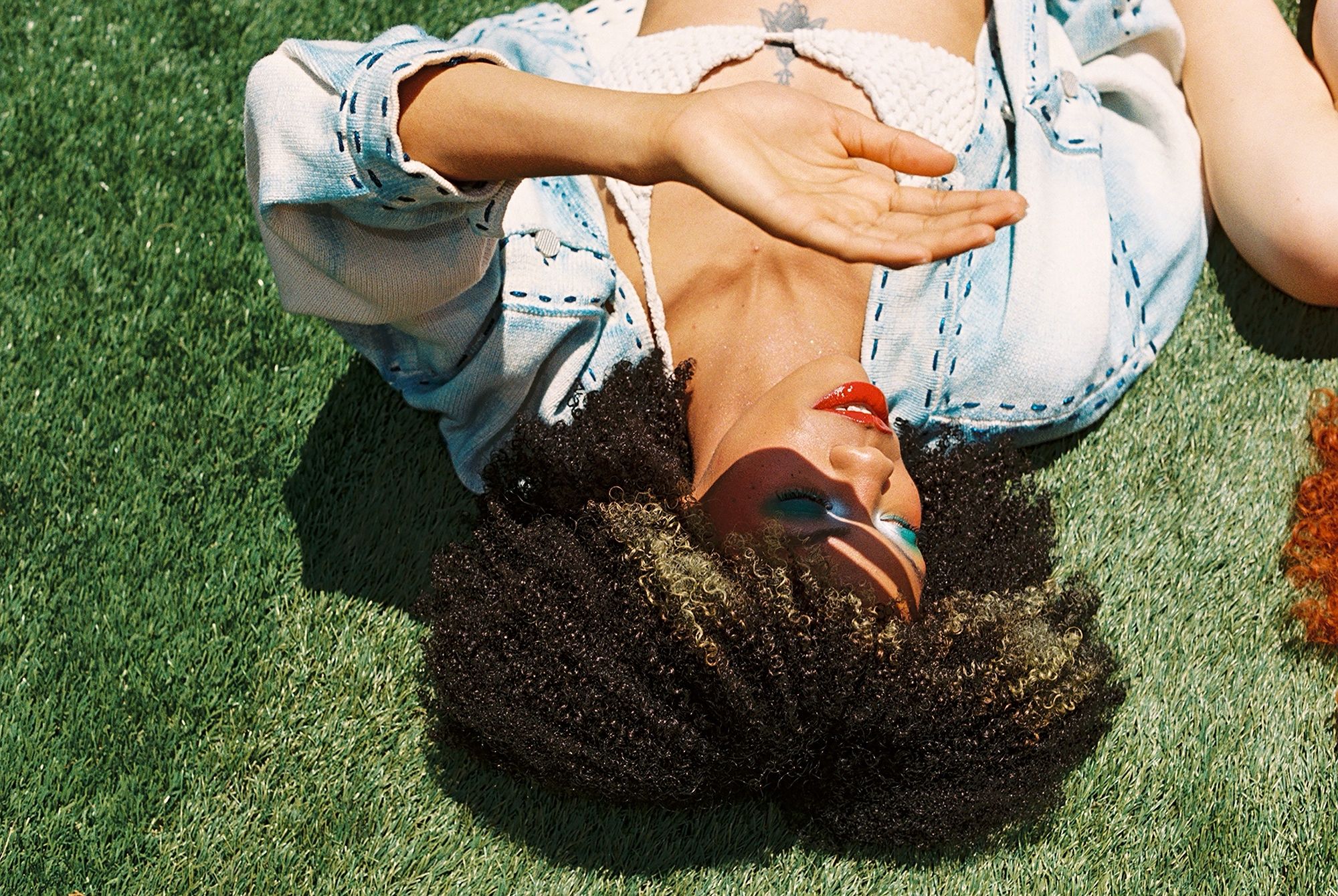 Upside-down close-up of a person lying on grass in a denim jacket, red lipstick and colorful eye makeup.