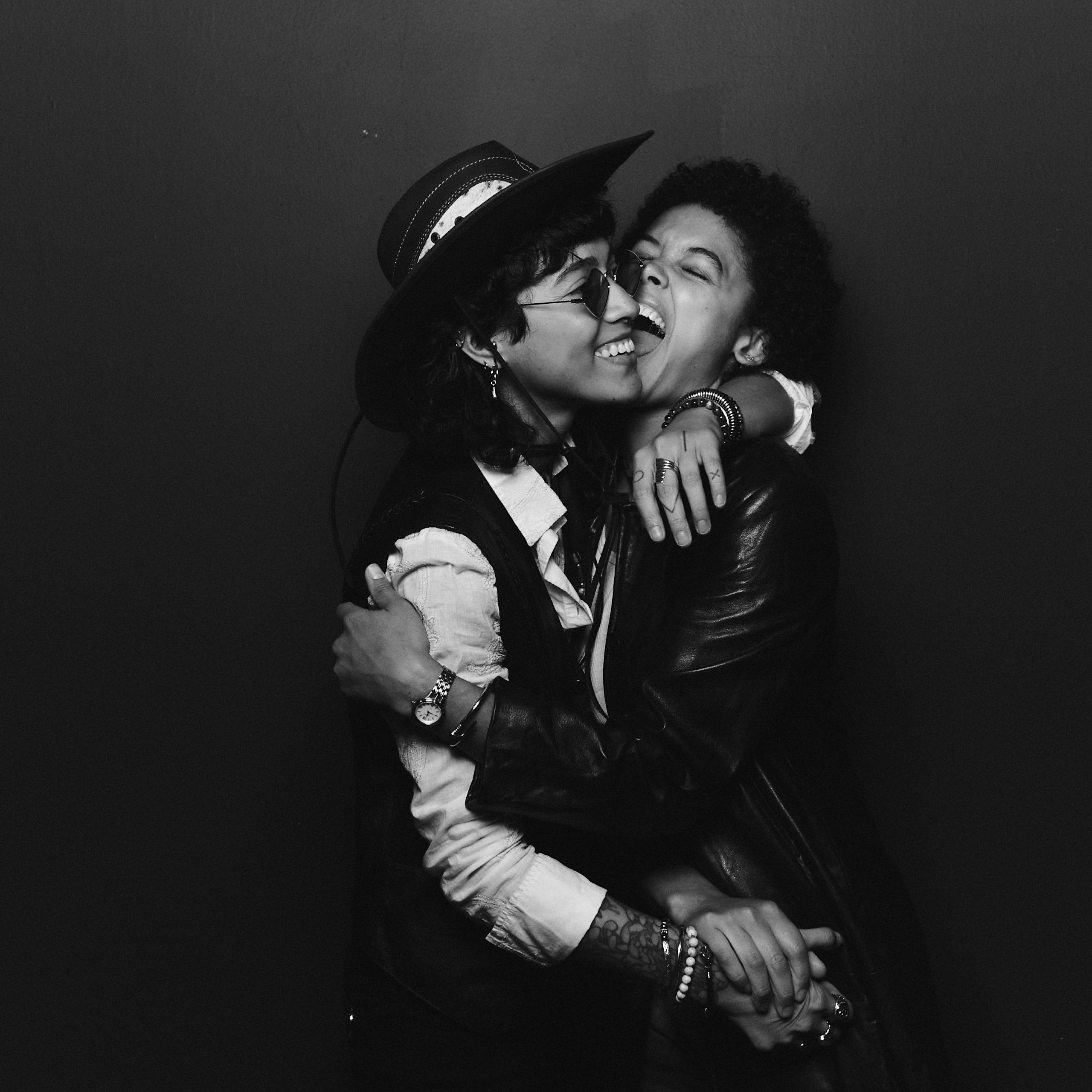 Black-and-white portrait of two people playfully embracing, one behind the other with arms around their shoulders.
