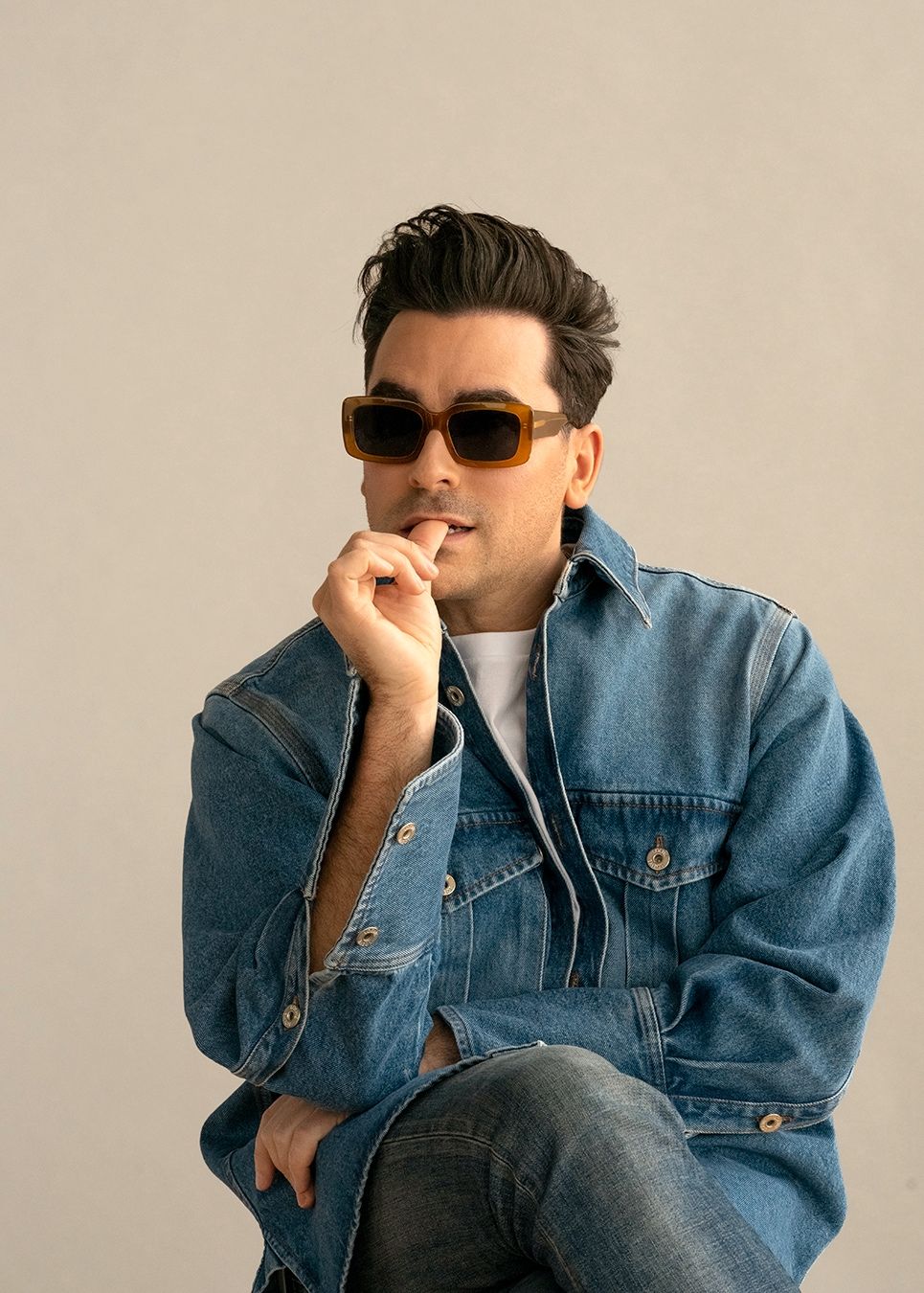 Man with styled hair and sunglasses in a denim jacket, hand raised near his mouth, studio portrait.