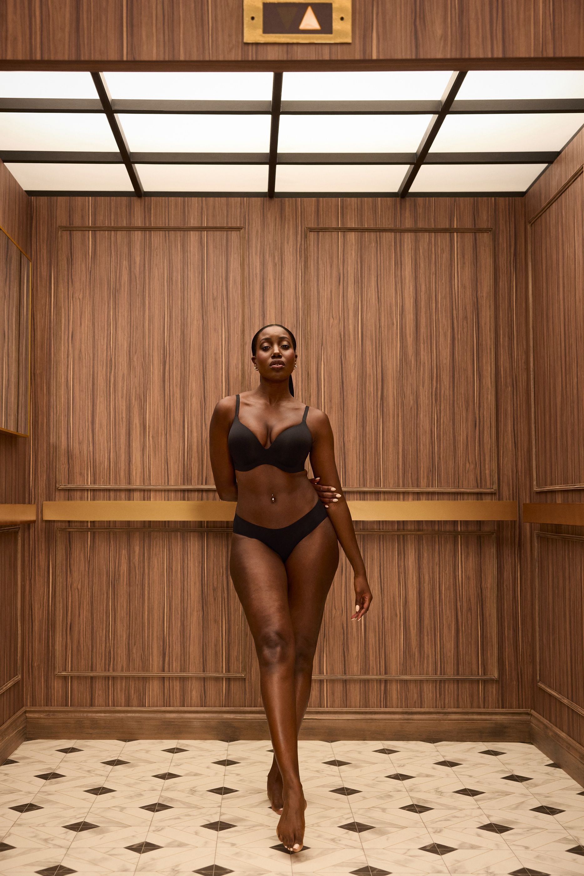 Woman in black lingerie stands in a wood-paneled elevator with patterned tile flooring.
