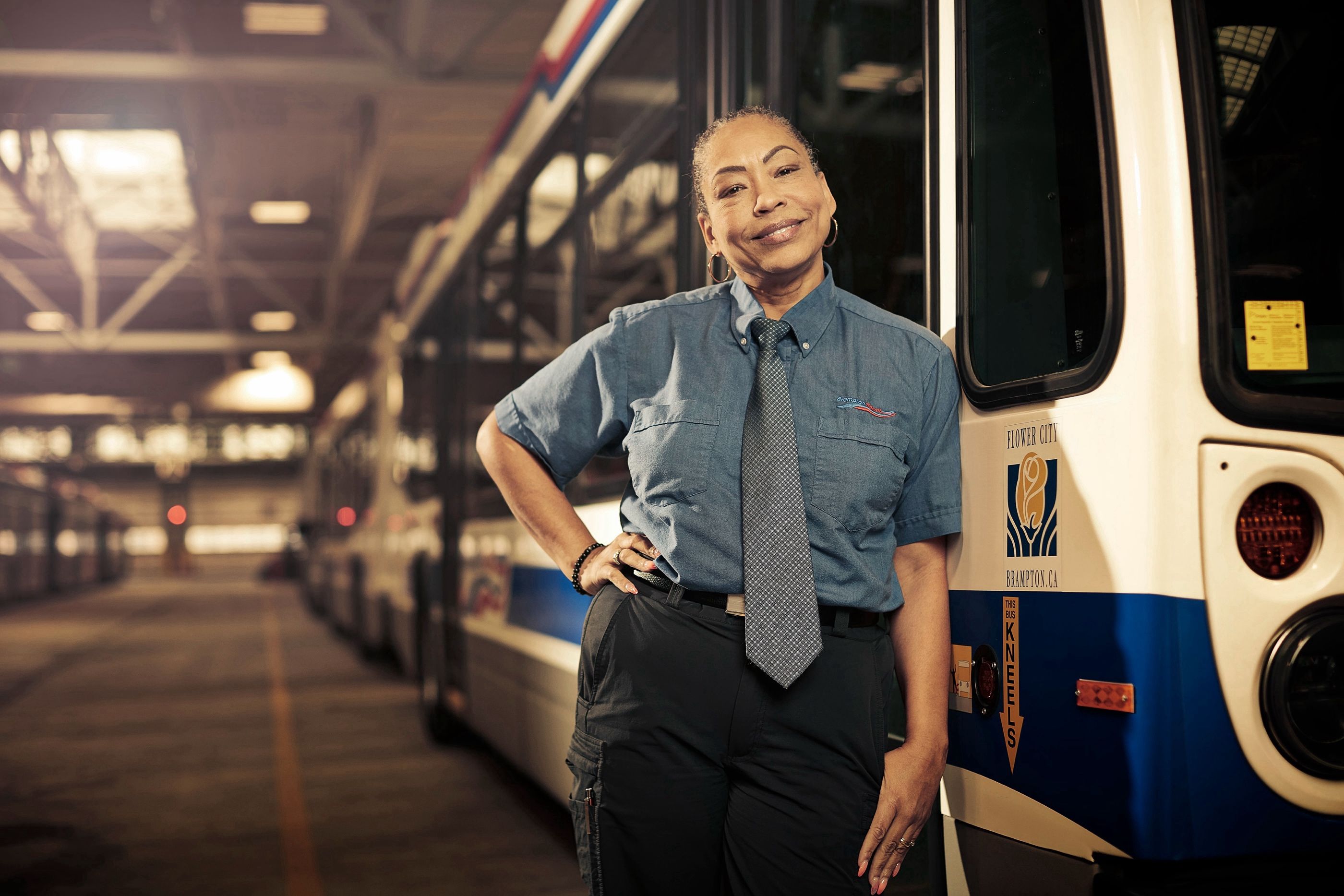 Woman in a transit uniform and tie stands beside a city transit bus in a garage, hand on hip, smiling.
