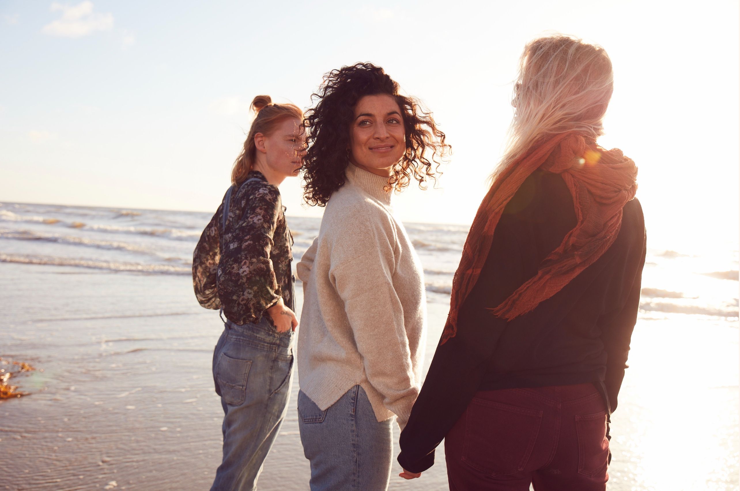 Three women walk along a beach at sunset, one glancing back over her shoulder.