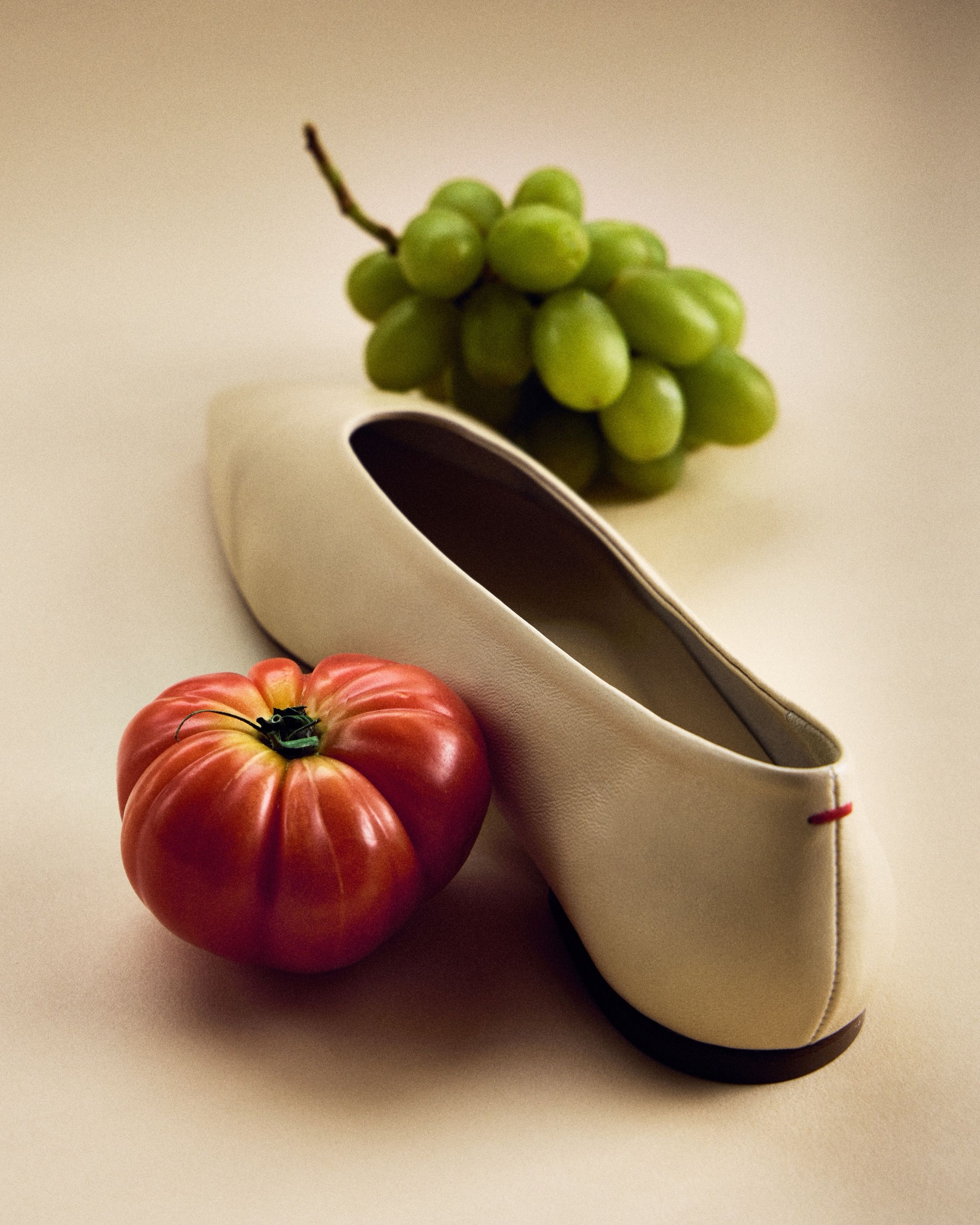 A cream leather flat shoe beside a heirloom tomato and a bunch of green grapes.