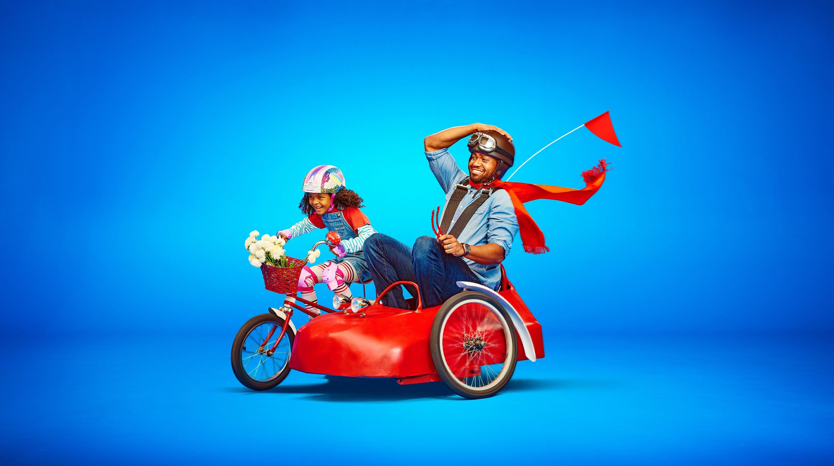 Man and girl ride a red bicycle with a sidecar against a solid blue background, both wearing helmets.