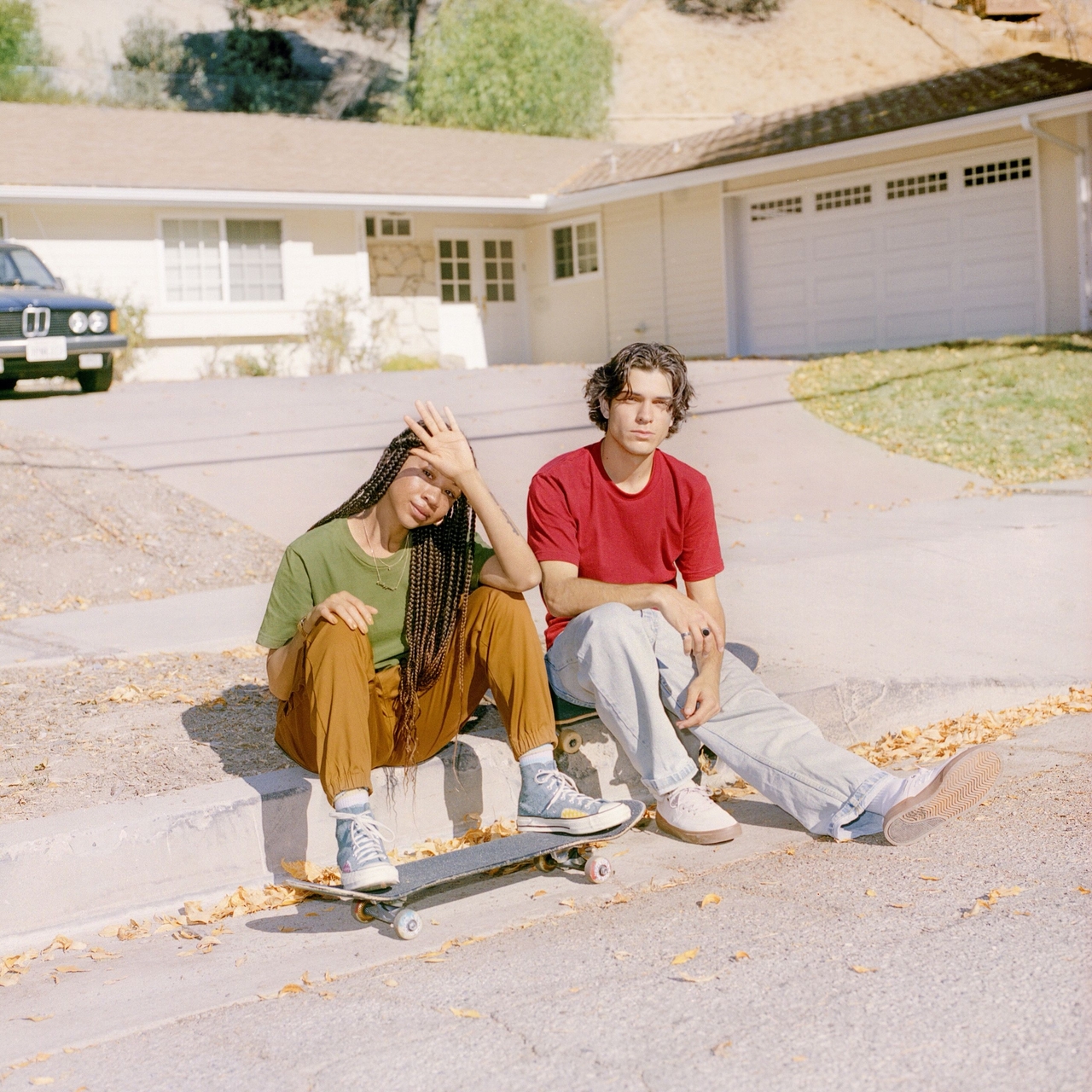 Two people sitting on a driveway curb next to a skateboard in front of a suburban house.