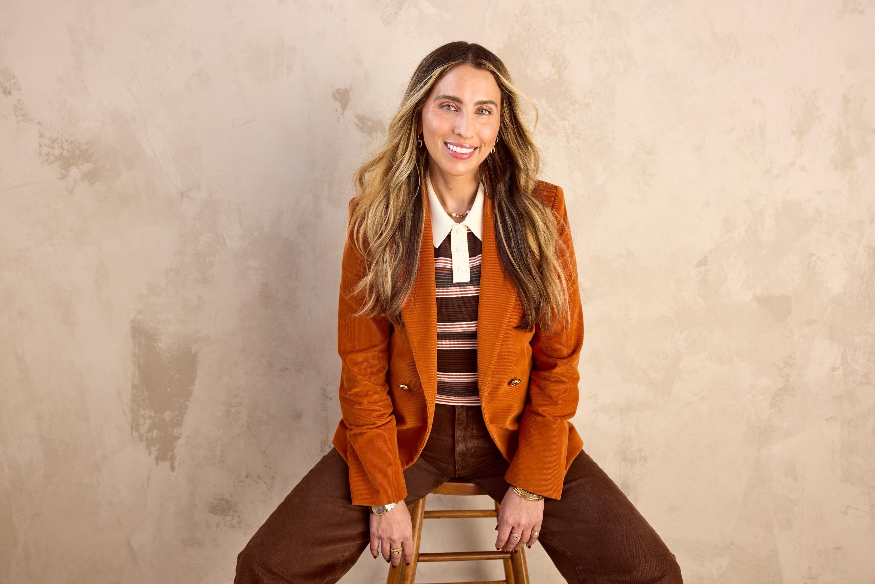 Woman in an orange corduroy blazer and striped top sitting on a wooden stool against a mottled beige backdrop.