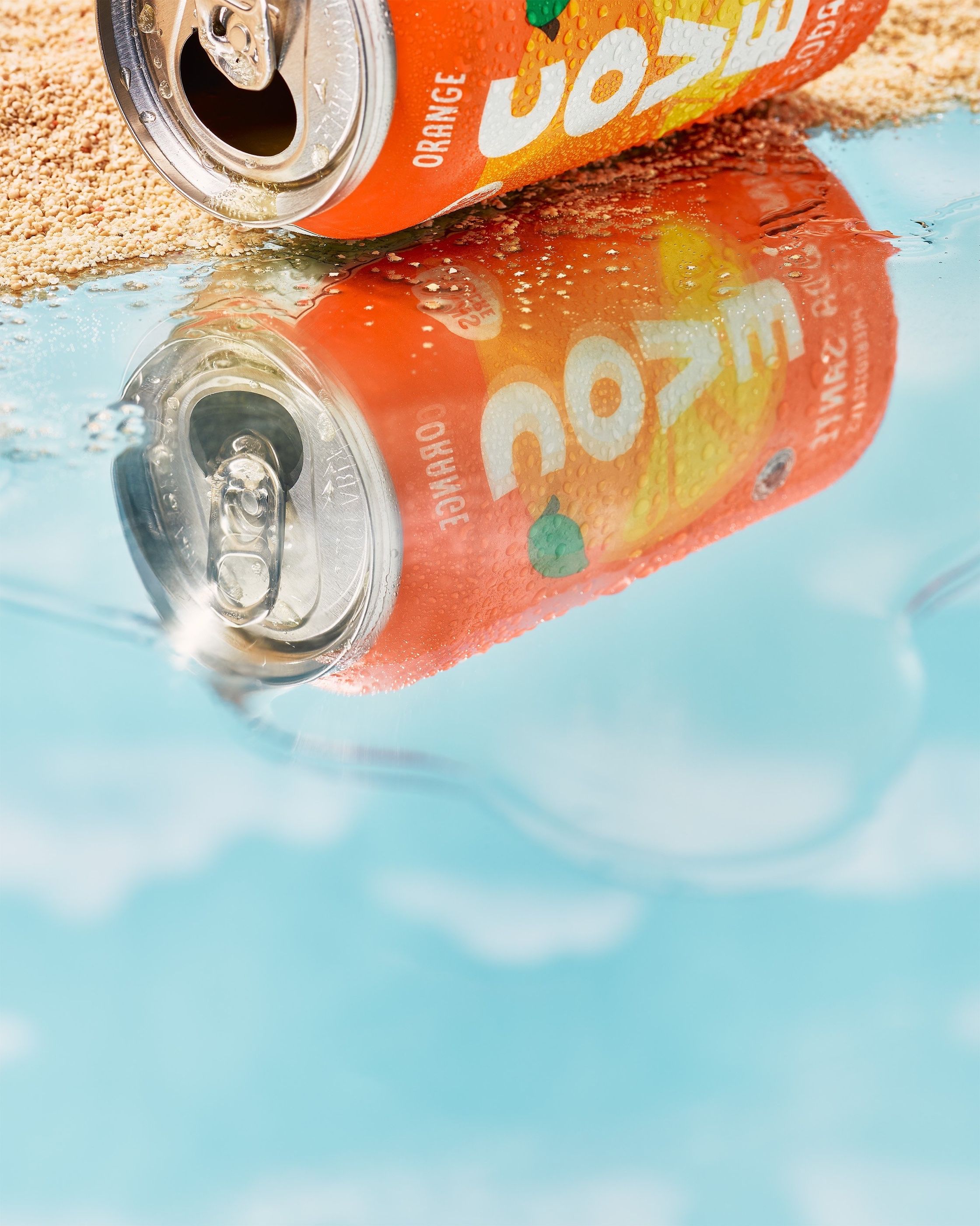 An orange soda can resting at the edge of water and sand, with its reflection visible on the wet surface.