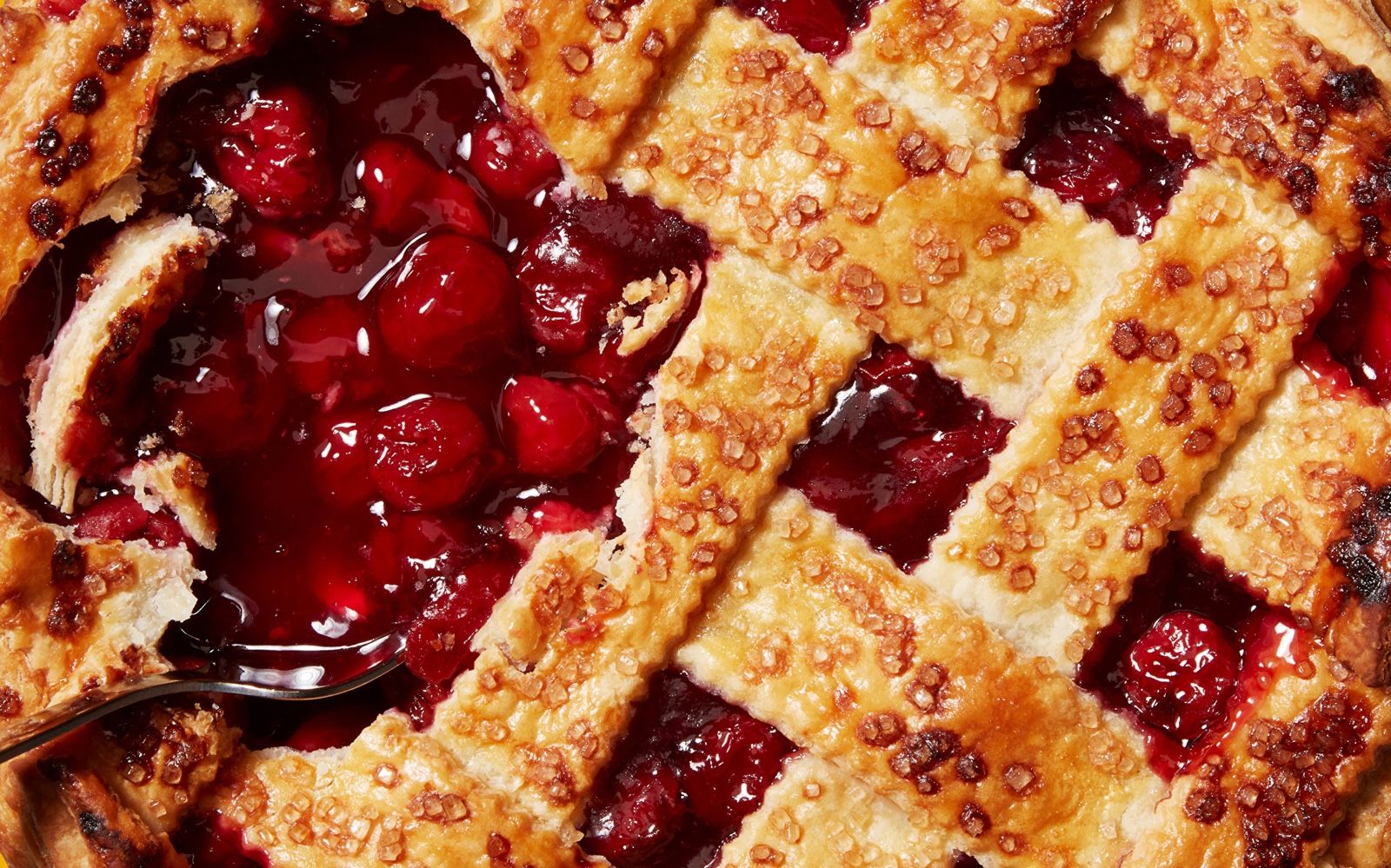 Close-up of a cherry pie with lattice crust, sugar crystals on top and a fork lifting the filling.