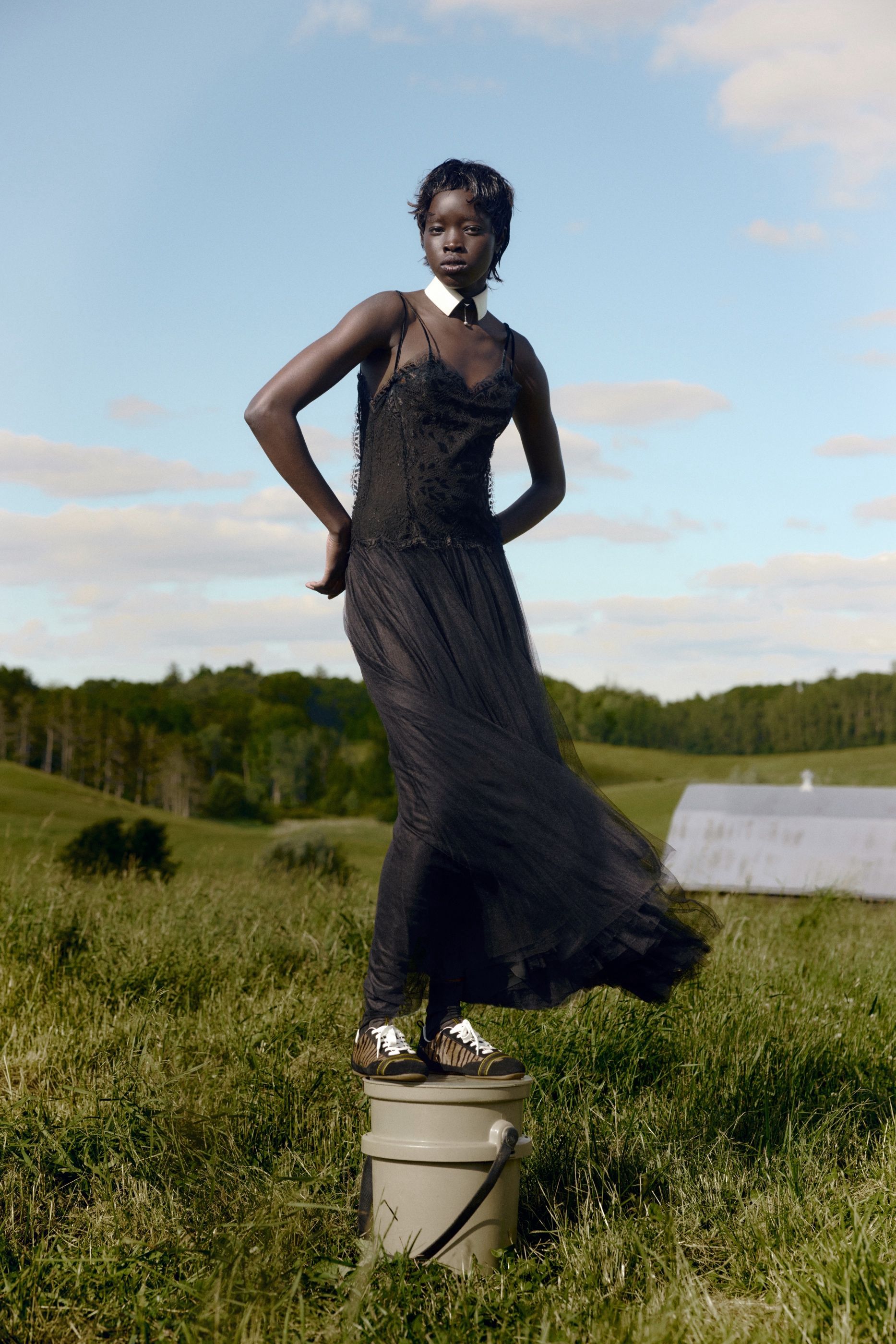 Woman in a black lace slip dress with a white collar and tie stands on a bucket in an open field, wearing striped sneakers.