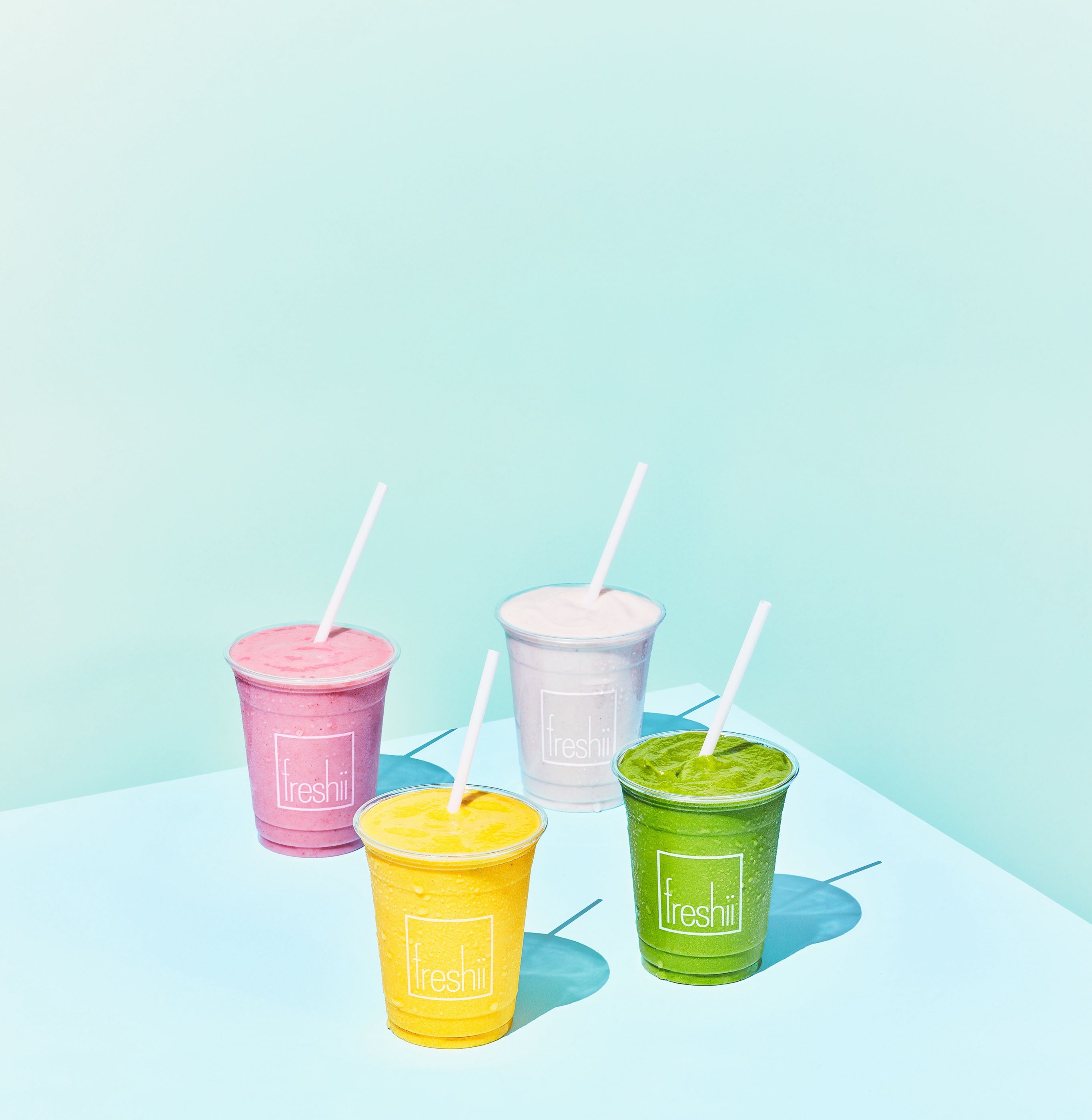 Four smoothies in Freshii-branded cups, in pink, yellow, white and green, arranged on a light teal surface.
