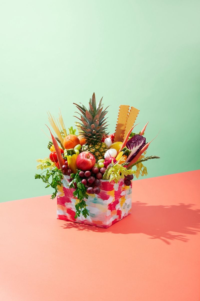 A patterned box overflowing with a pineapple, grapes, asparagus, radishes, carrots and other fresh produce.