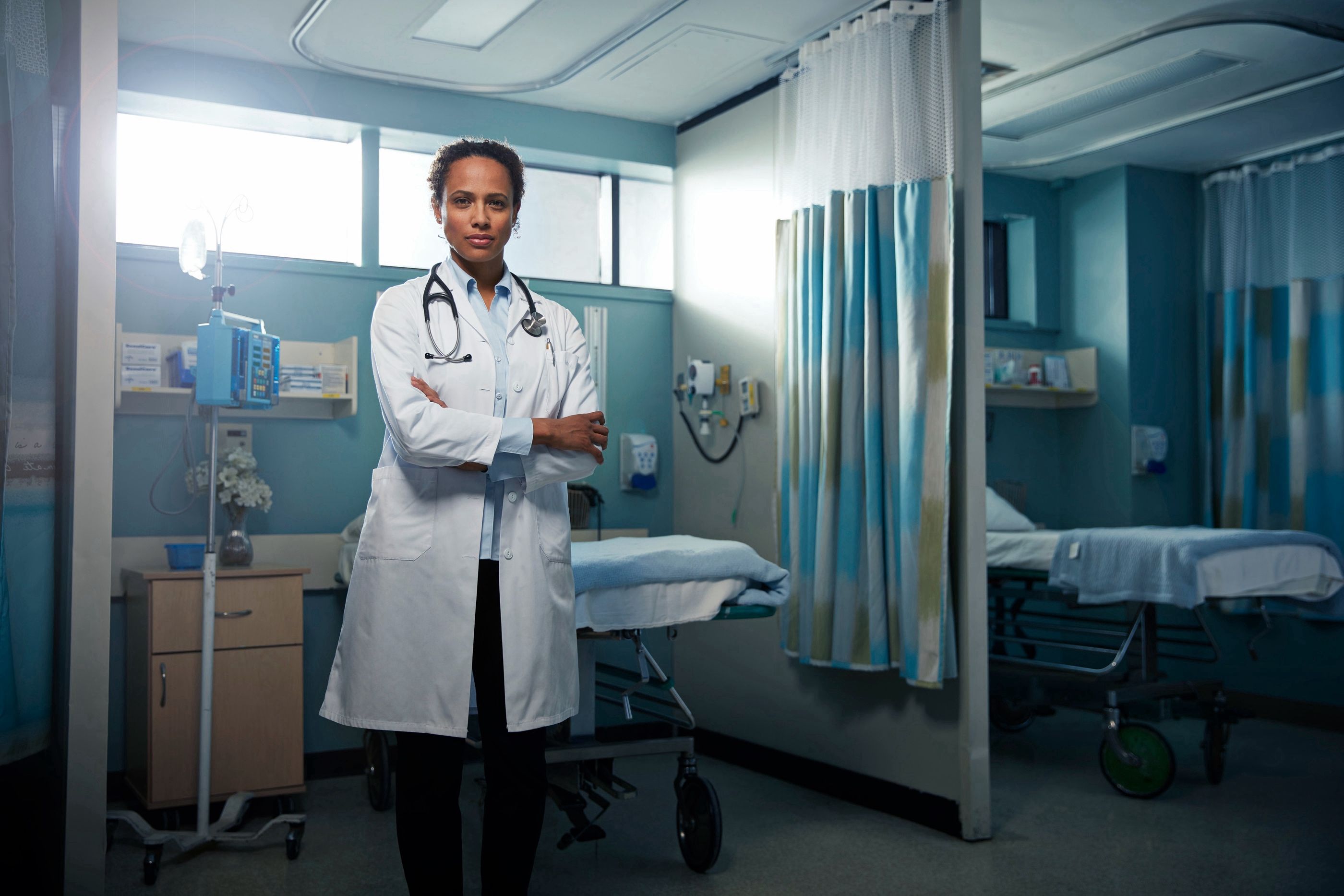 Doctor in a white coat and stethoscope stands with arms crossed in an empty hospital patient room.