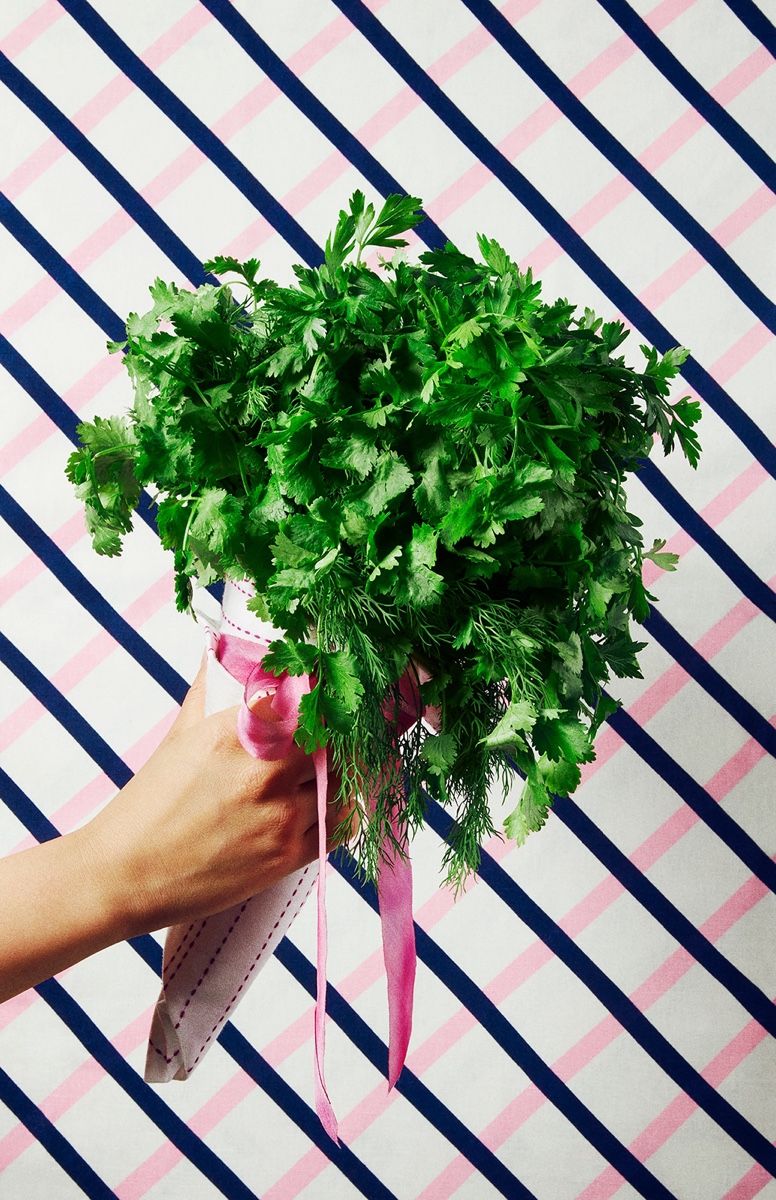 A hand holds a bunch of fresh cilantro wrapped in paper with a pink ribbon, against a striped background.